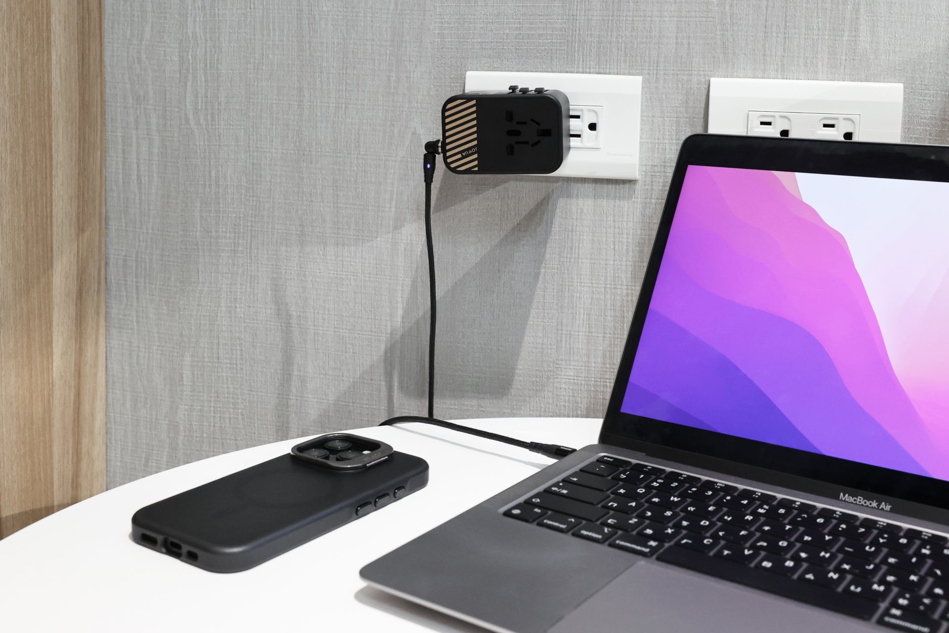 On a white table, a laptop with a vibrant display is connected to the wall outlet through the JOWUA 65W GaN Travel Adapter. Nearby, a smartphone enjoys the benefits of fast charging power from this advanced technology, all set against the wall's calm neutral texture.