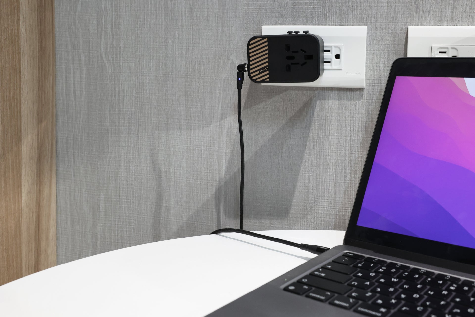 A laptop on a white table is connected to a JOWUA 65W GaN Travel Adapter plugged into a wall socket. The adapter, with its wood texture, supports 65W Fast Charging via the USB cable extending to the laptop, against a gray textured wall.