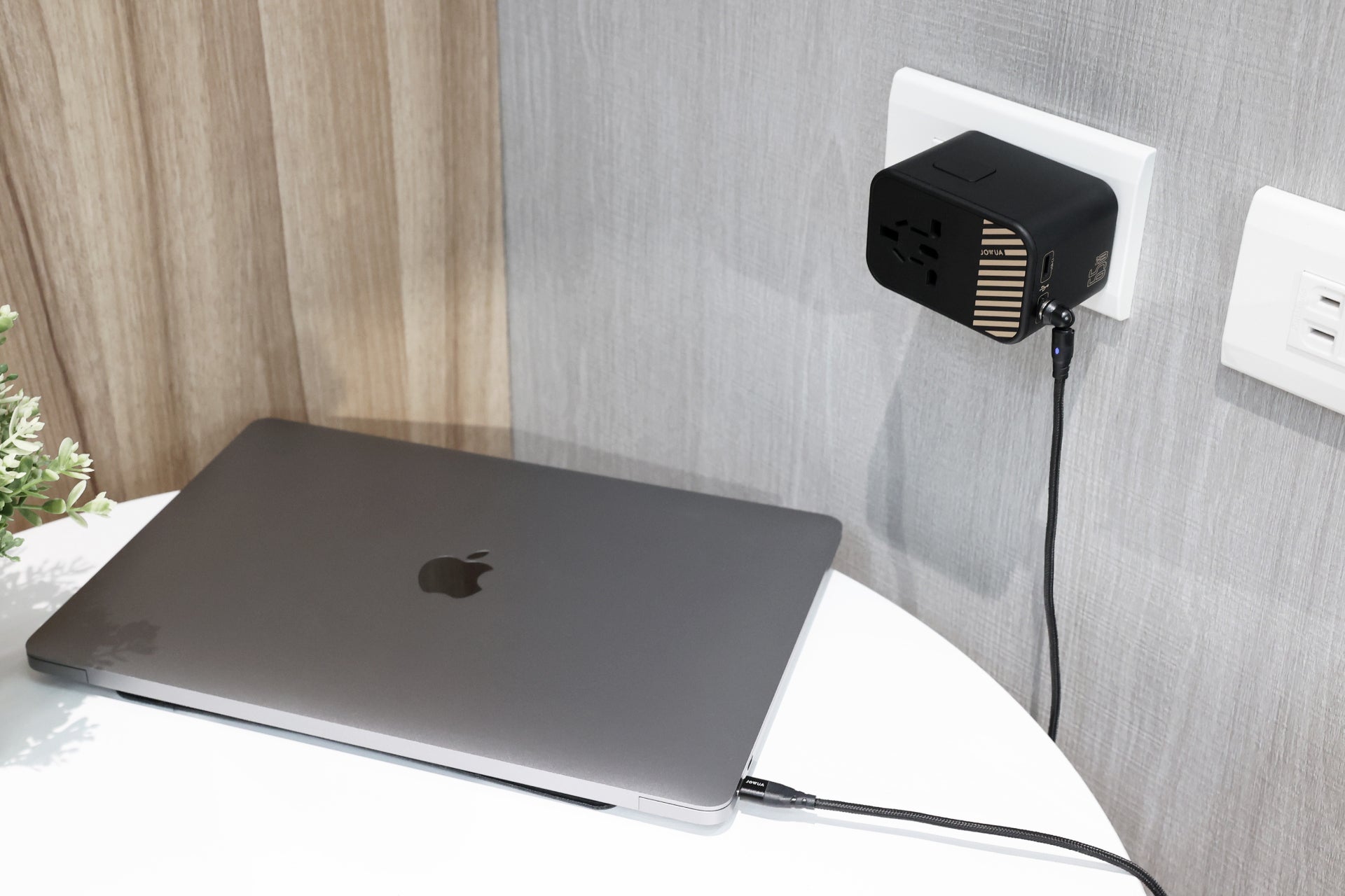 A silver laptop sits on a white round table, connected to the wall socket through a JOWUA 65W GaN Travel Adapter in black. This adapter supports international charging and includes multiple plug styles. The wall showcases wooden panels with grey wallpaper, and part of a small potted plant is visible.