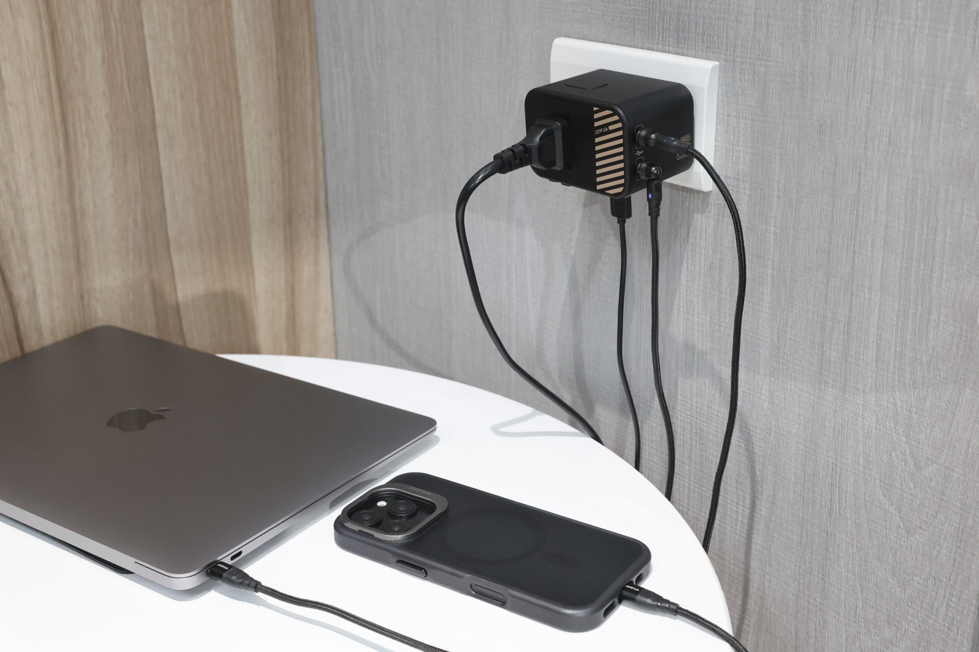 A laptop and smartphone are charging on a small white table, powered by a sleek black JOWUA 65W GaN Travel Adapter. Multiple cables extend from it to the wall socket, ensuring efficient fast charging. The scene is set against light-colored wood and gray walls.