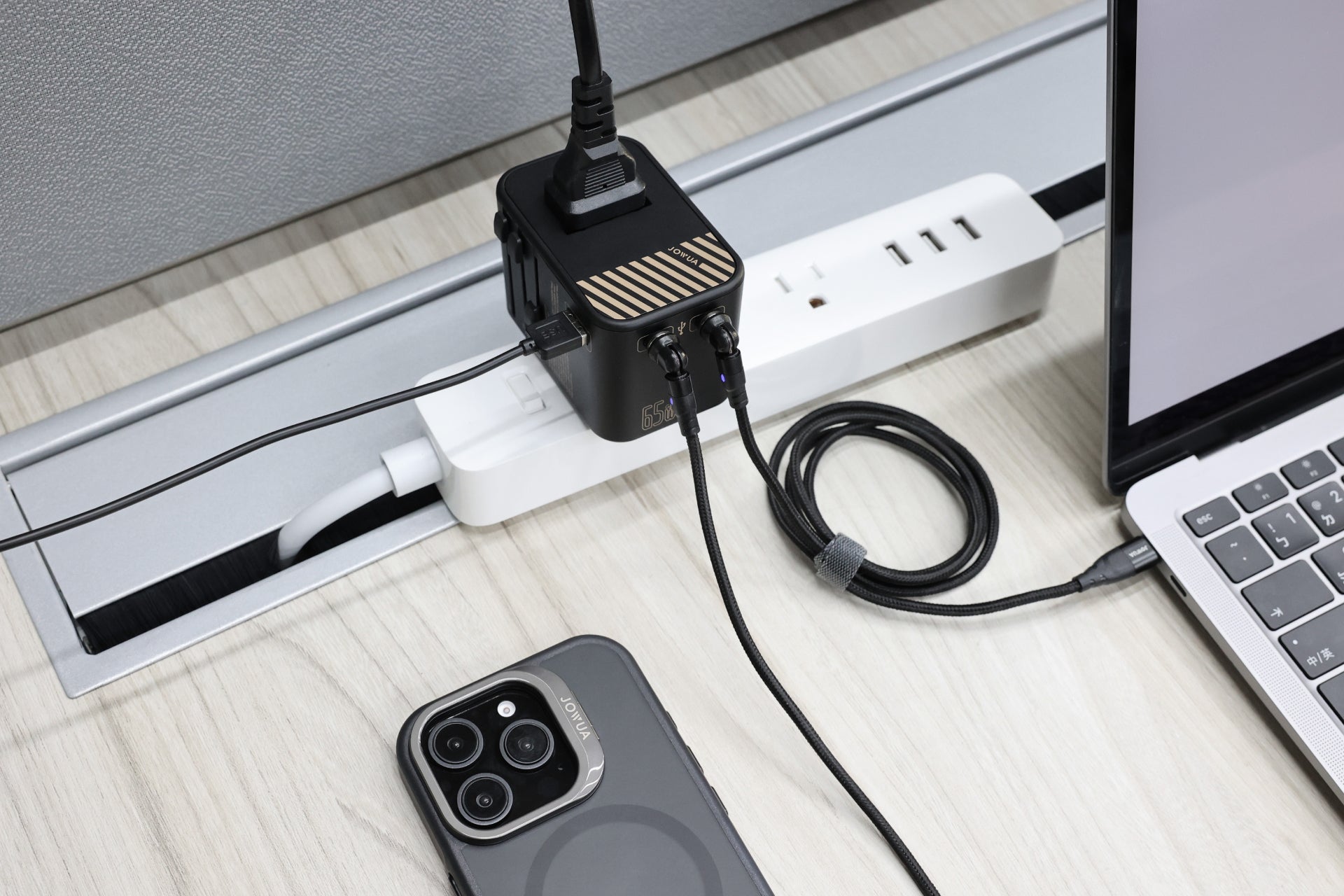 A smartphone and laptop are connected to a JOWUA 65W GaN Travel Adapter on a desk, utilizing the high-speed charging feature. A charging cable powers the phone, while another energizes the laptop.