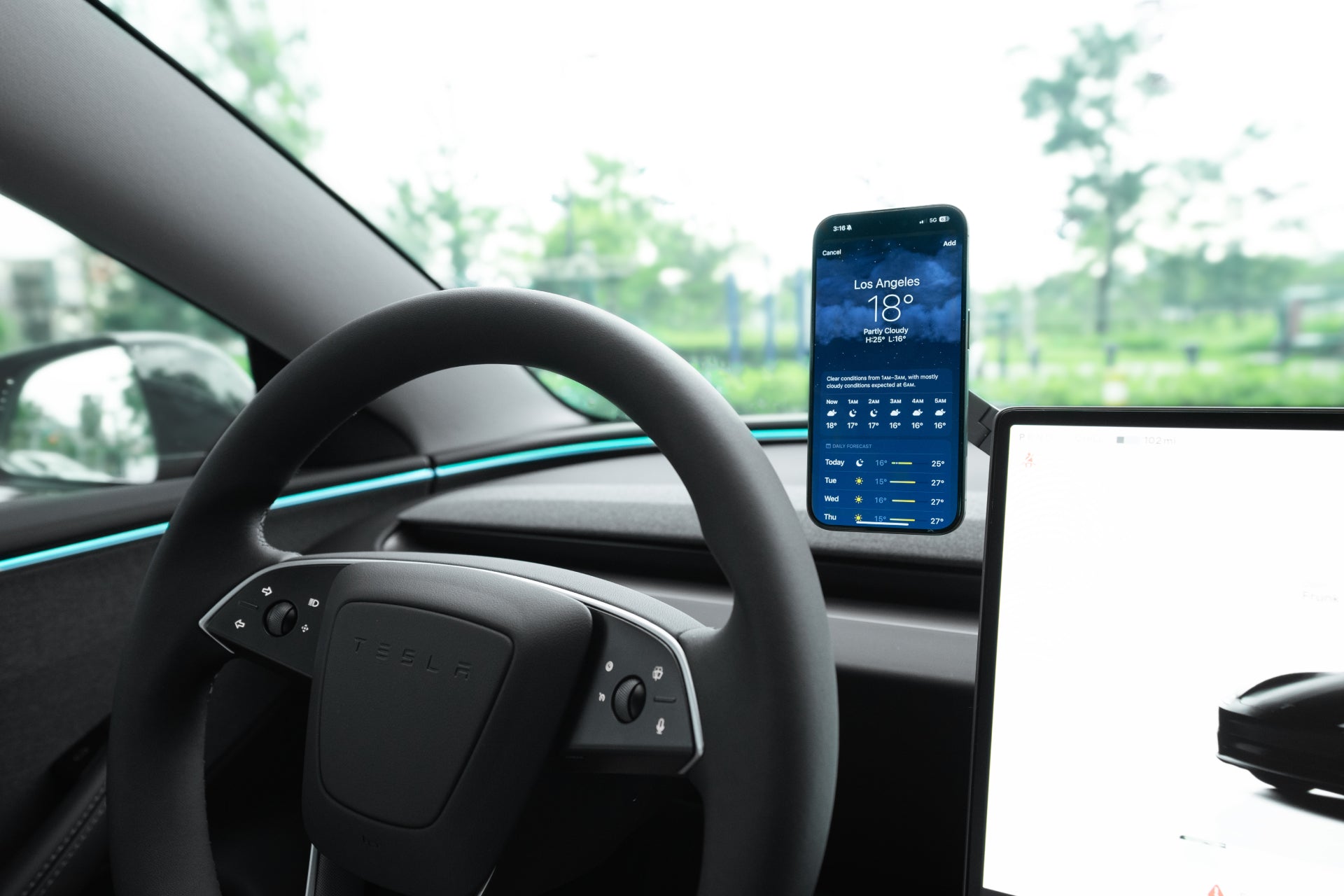 Interior view of a car highlighting the Tesla Christmas : The Dream Team by JOWUA, featuring a sleek steering wheel and a smartphone displaying a weather app for Los Angeles at 18°C, securely held in place by a MagSafe phone mount. The dashboard screen showcases an image of the car, with vibrant greenery visible through the windows in the background.