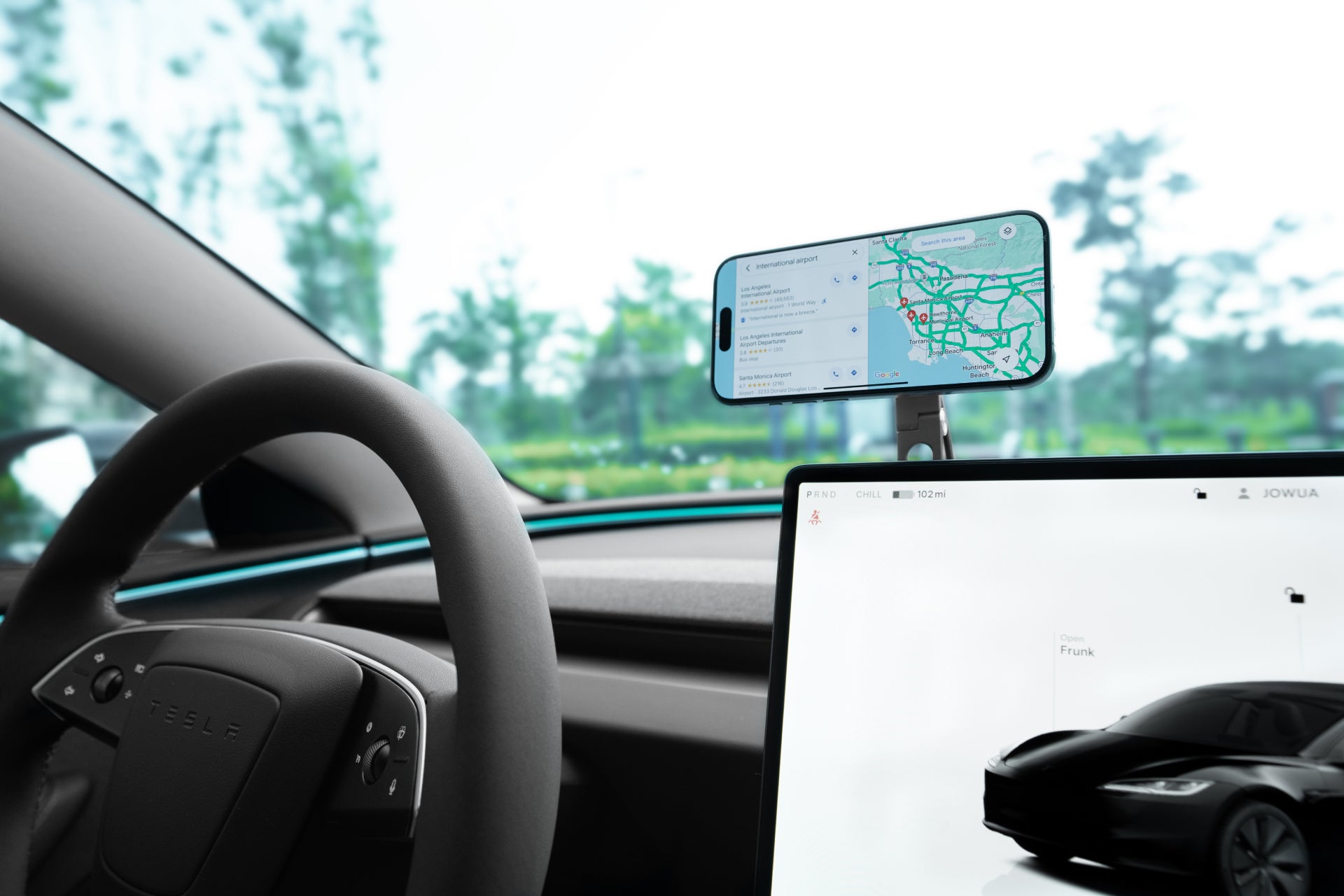 The interior of a Tesla Model 3 Highland features a steering wheel and a JOWUA 6D Tesla Compatible Invisible Foldaway Car Mount Qi2 Wireless Charger, securely holding a smartphone with MagSafe wireless charging, displaying map directions. A built-in touchscreen showcases a car image, with blurred greenery visible through the windows.