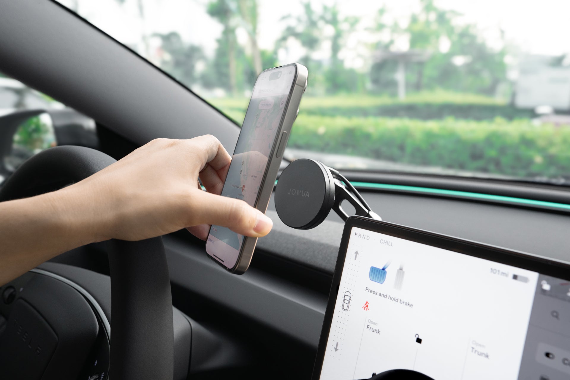 A person installs a JOWUA Tesla Christmas: The Dream Team phone mount inside their vehicle, effortlessly securing their smartphone while presenting the map. Meanwhile, the dashboard screen displays car settings, and the backdrop features a blurred view of greenery outside the window, emphasizing the convenience of this Tesla accessory.