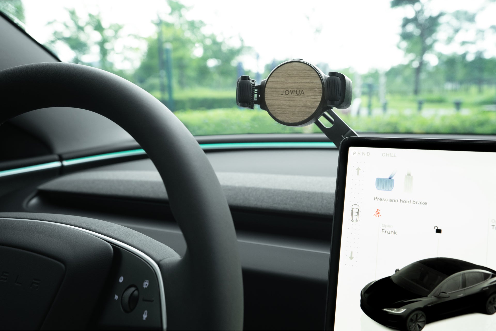 The car interior close-up features a steering wheel, digital display with vehicle diagram, and a JOWUA 6D Tesla Compatible Invisible Foldaway Car Mount Qi2 Wireless Charger attached to the dashboard. Through the windshield, there's a blurred green landscape.