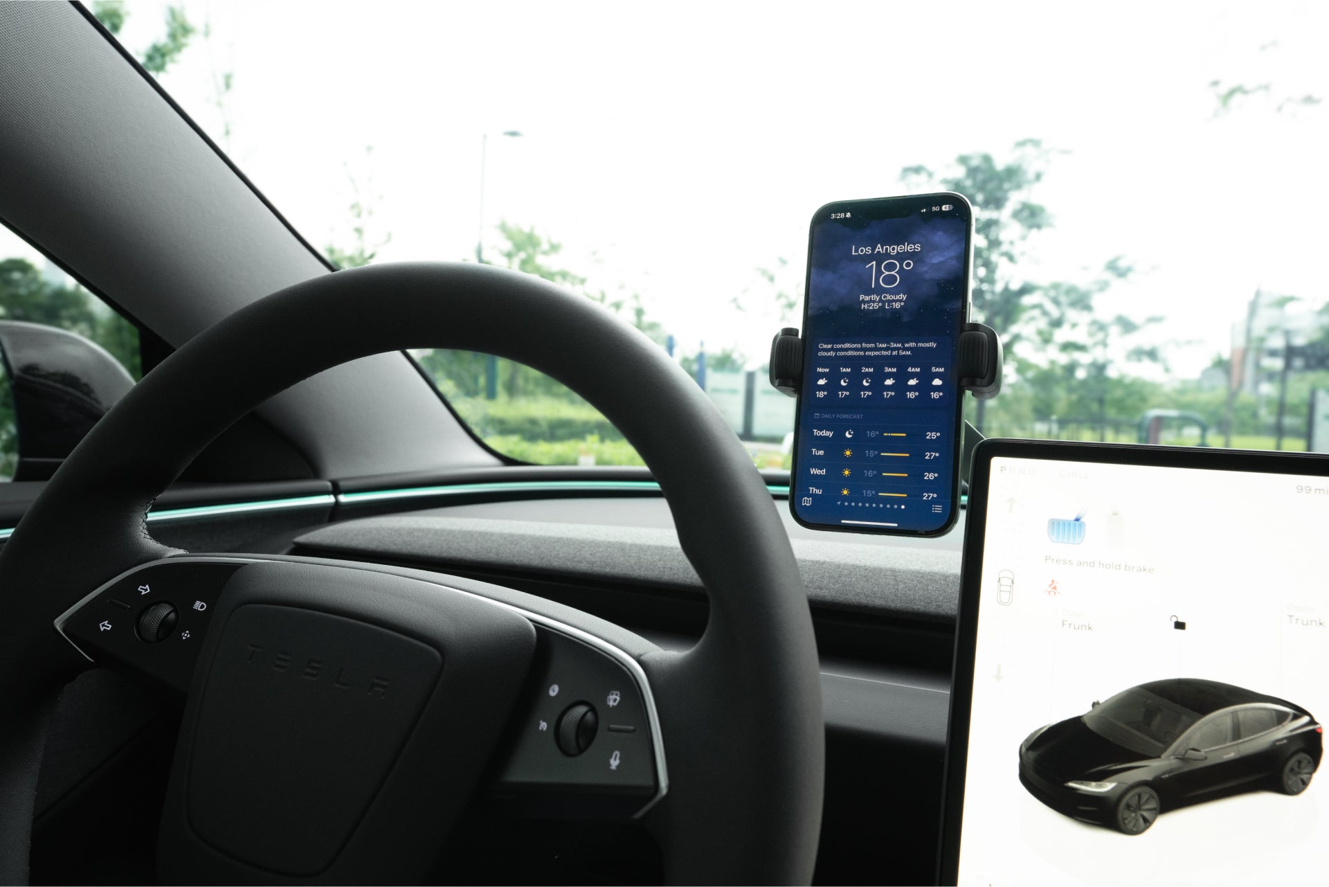 A JOWUA 6D Tesla Compatible Invisible Foldaway Car Mount Qi2 Wireless Charger secures a smartphone on the steering wheel and dashboard of a Tesla, displaying Los Angeles weather. The touchscreen shows vehicle status, while greenery is visible through the windows.