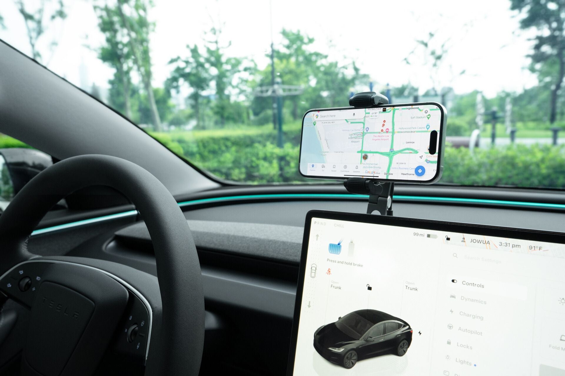 Inside the car, a digital dashboard displays a sleek black model, while a smartphone on JOWUA's 6D Tesla Compatible Foldaway Car Mount Qi2 Wireless Charger features navigation. Through the windshield, lush green trees and a cloudy sky create a tranquil backdrop.