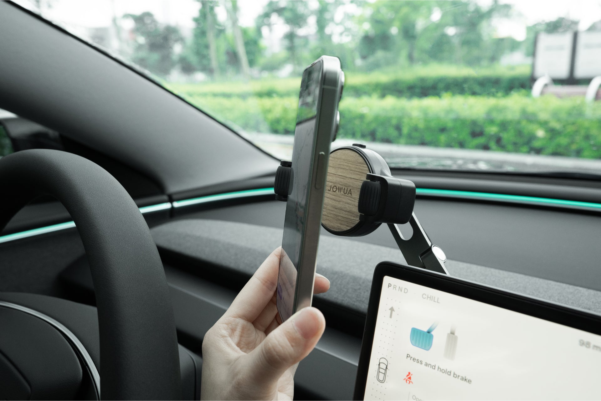 A person secures a smartphone into the JOWUA 6D Tesla Compatible Invisible Foldaway Car Mount Qi2 Wireless Charger on a Model 3 dashboard. The interior and steering wheel are visible, along with greenery outside, while the screen displays controls and highlights the Qi2 charging feature.