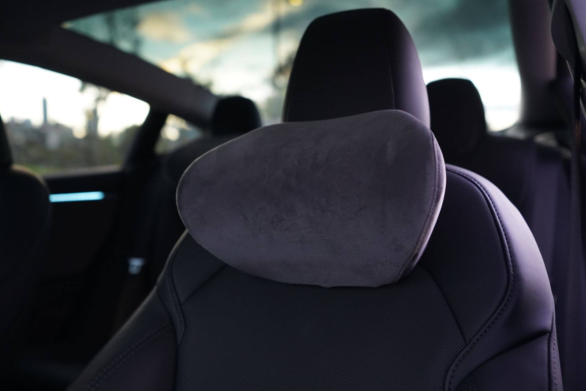 Close-up view of a JOWUA Car Neck Pillow with an ergonomic design and plush headrest cover in a dimly lit interior, showcasing the texture and comfort. The window in the background reveals a blurry landscape outside.