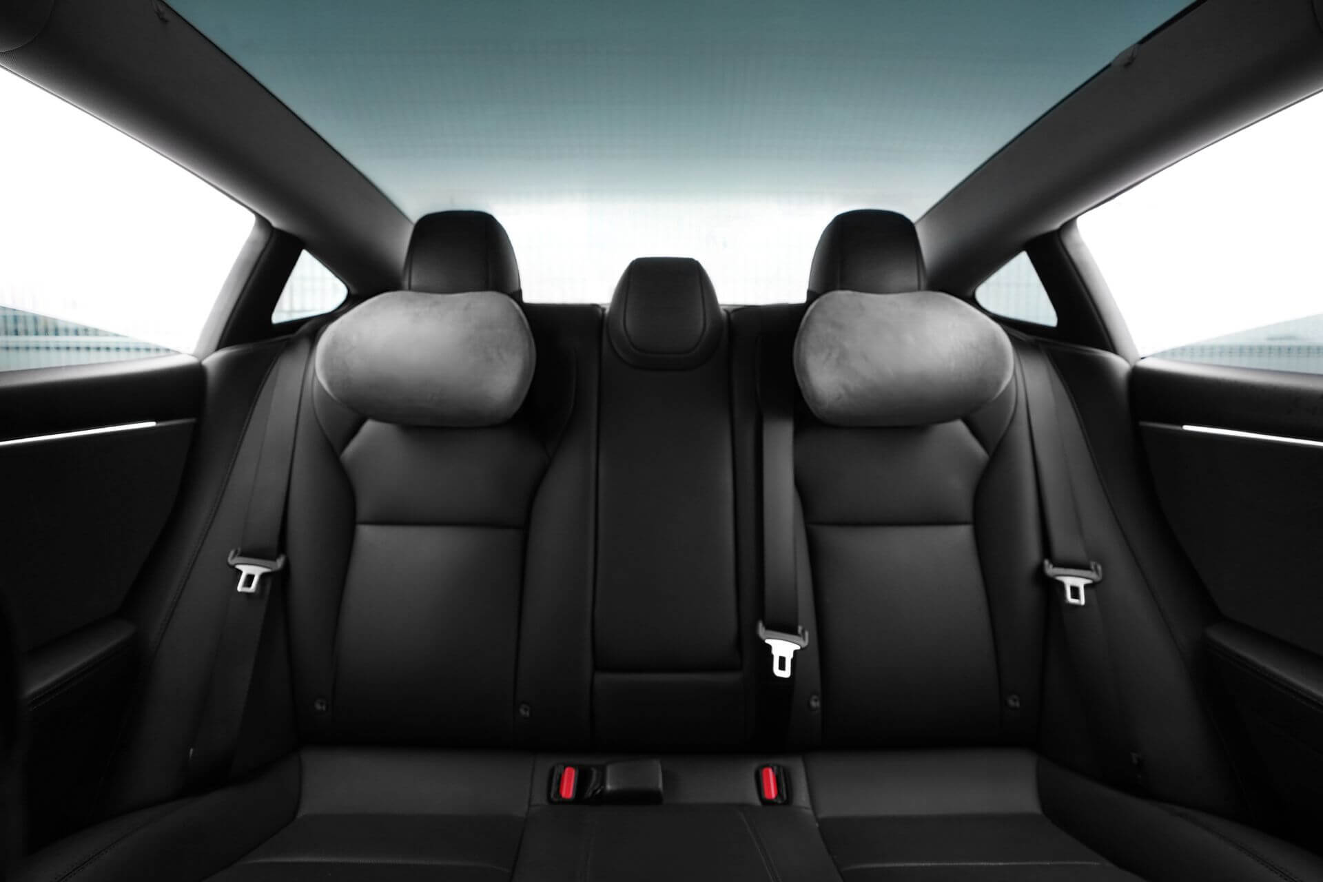 Discover the interior of a contemporary car's backseat, highlighted by sleek black leather upholstery and two mounted seatbelt buckles. Soft gray JOWUA Car Neck Pillows rest on the adjustable headrests for enhanced driving comfort, while a sunroof allows light to filter through.