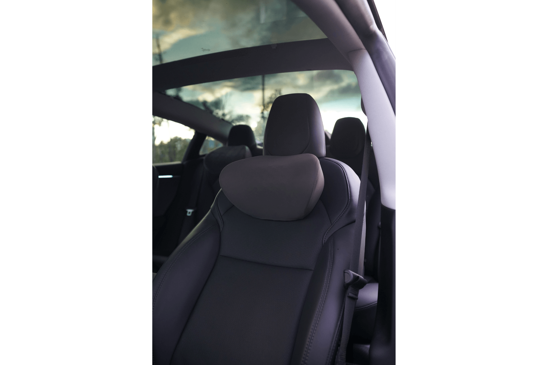 Interior view of a car highlighting a close-up of the front passenger seat with a JOWUA Car Neck Pillow. The black leather seat, designed ergonomically, sits beneath an expansive panoramic glass roof, through which a slightly cloudy sky is visible. The angle suggests a spacious and modern aesthetic.