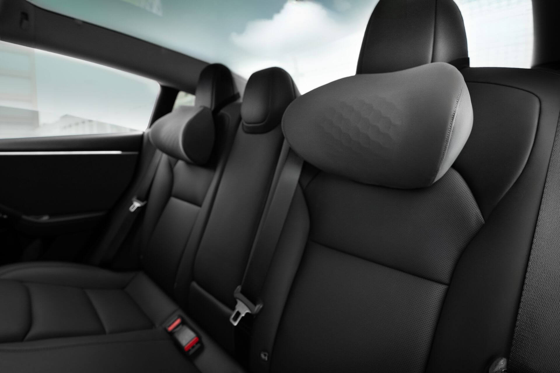 Inside the car, three ergonomically crafted black leather seats and two seat belts are visible. The window behind blurs a building view, while the headrests feature a textured pattern in dual materials for comfort. Enhance your ride with the JOWUA Car Neck Pillow for added support.