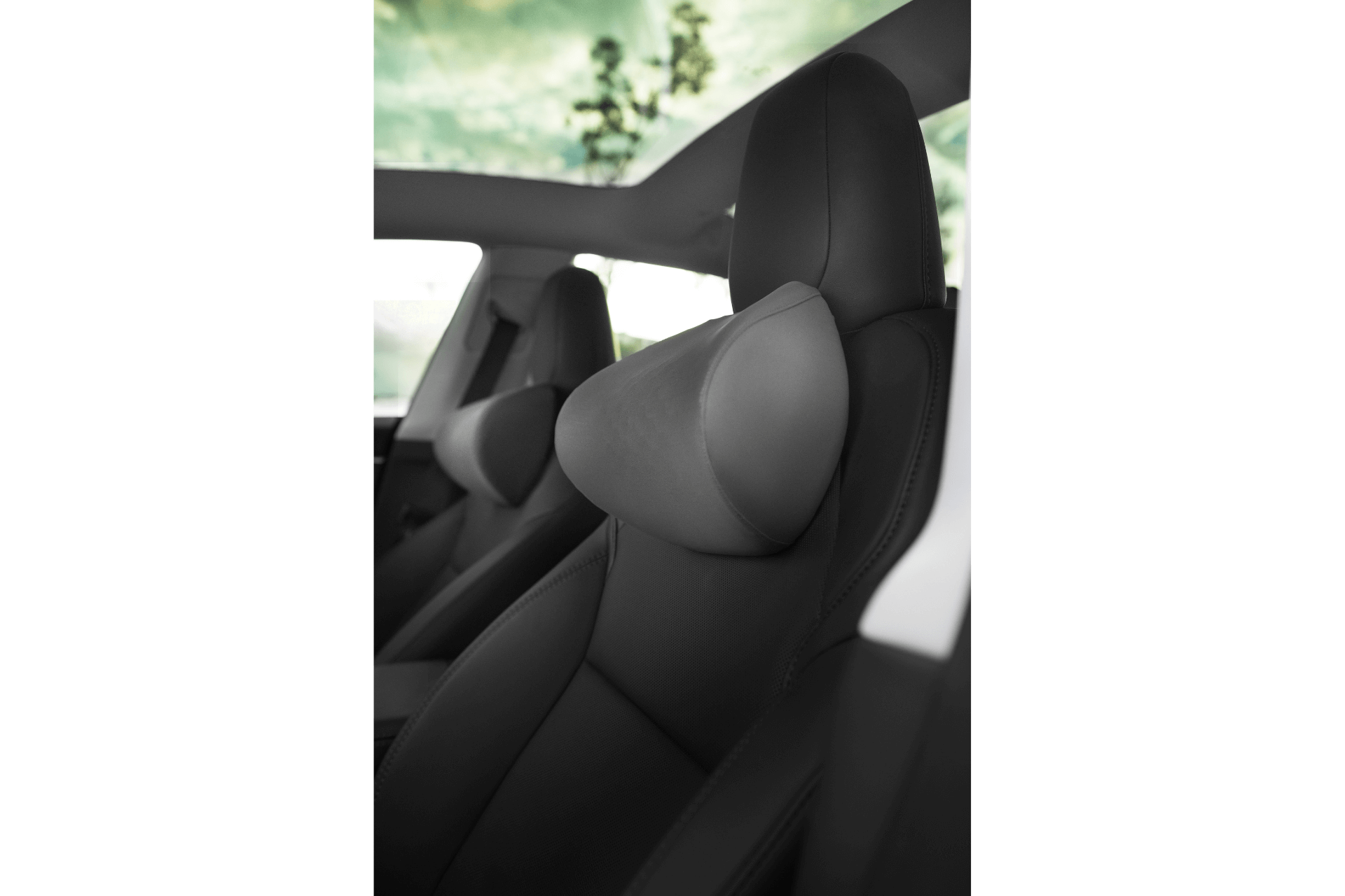 Interior view of a car featuring sleek black leather seats with a JOWUA Car Neck Pillow. Sunlight filters through the panoramic sunroof, highlighting hints of the partly cloudy green sky visible through the windows and enhancing the dual-material combination interior.