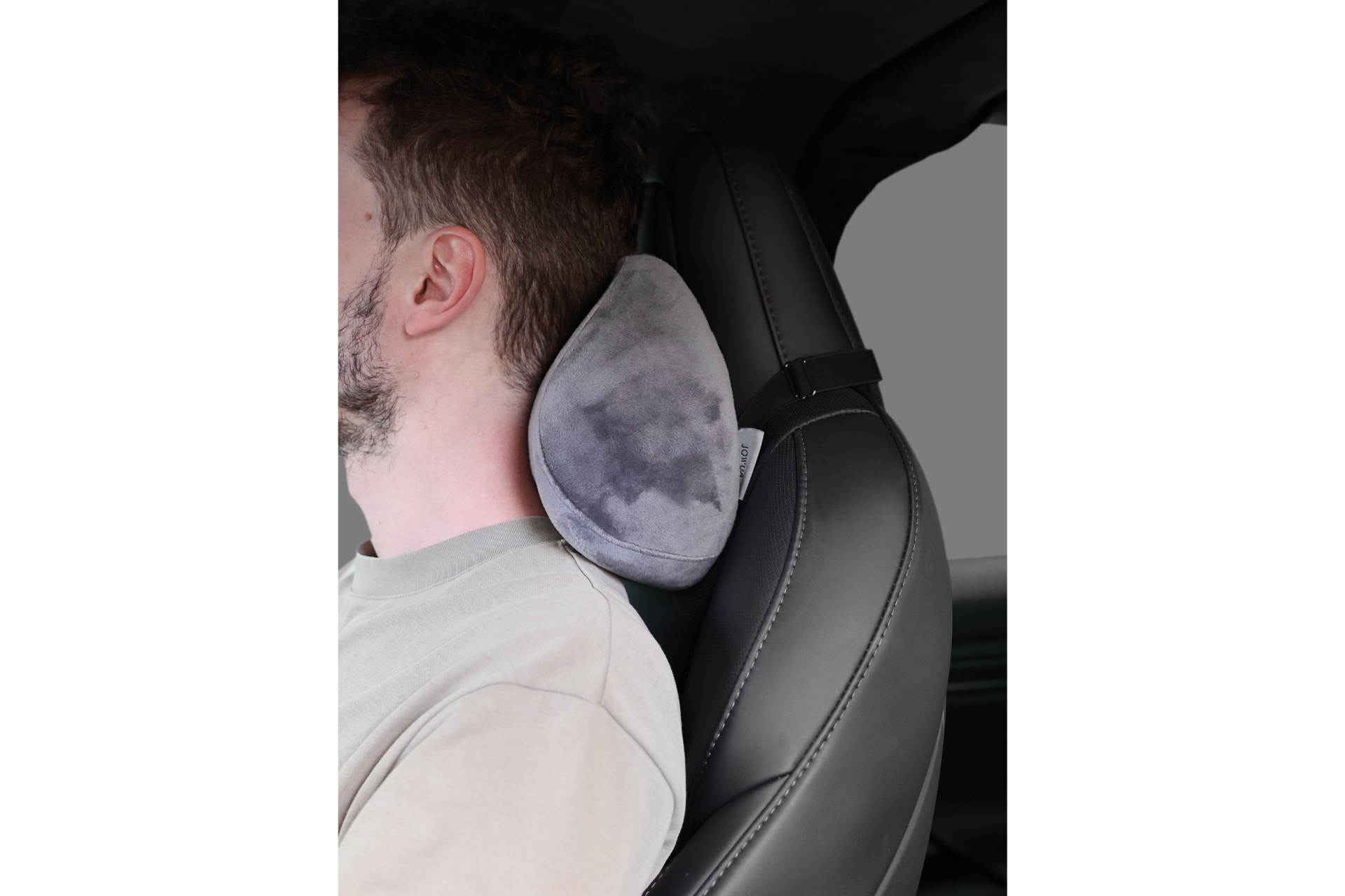 A man rests his head on a JOWUA Car Neck Pillow, made of gray memory foam with an ergonomic design, attached to the car's headrest for support and comfort while driving.