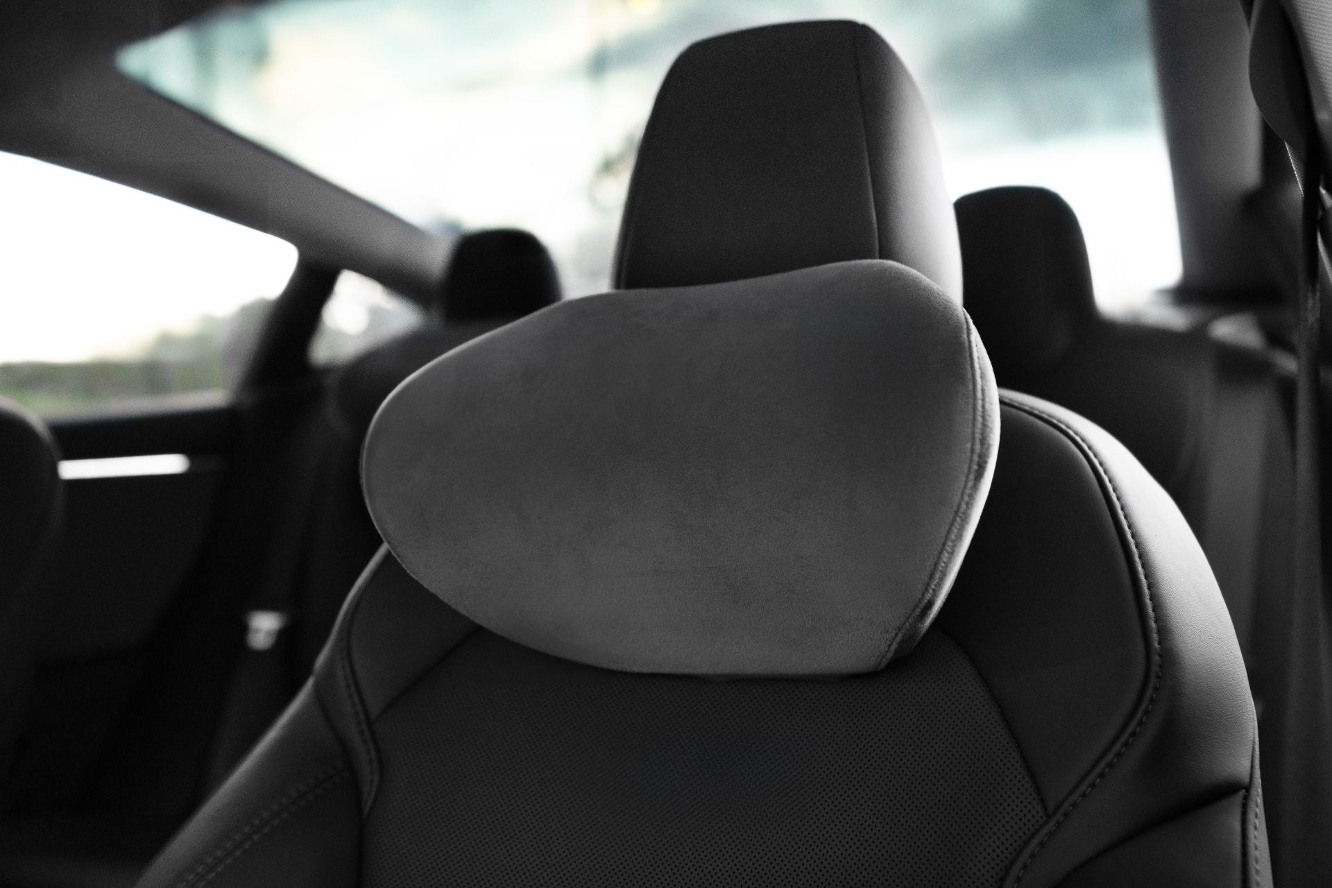 Close-up of a JOWUA Car Neck Pillow on an ergonomic seat with a dark, textured headrest. The sleek interior is softly illuminated by sunlight, with a blurred window in the background.