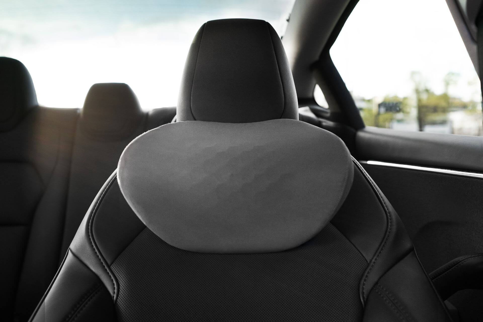 Close-up of a JOWUA Car Neck Pillow in a black, ergonomic design complementing the front seat's cushioned headrest. The smooth texture enhances comfort. Blurred rear seats and windows in the background suggest daylight outside.