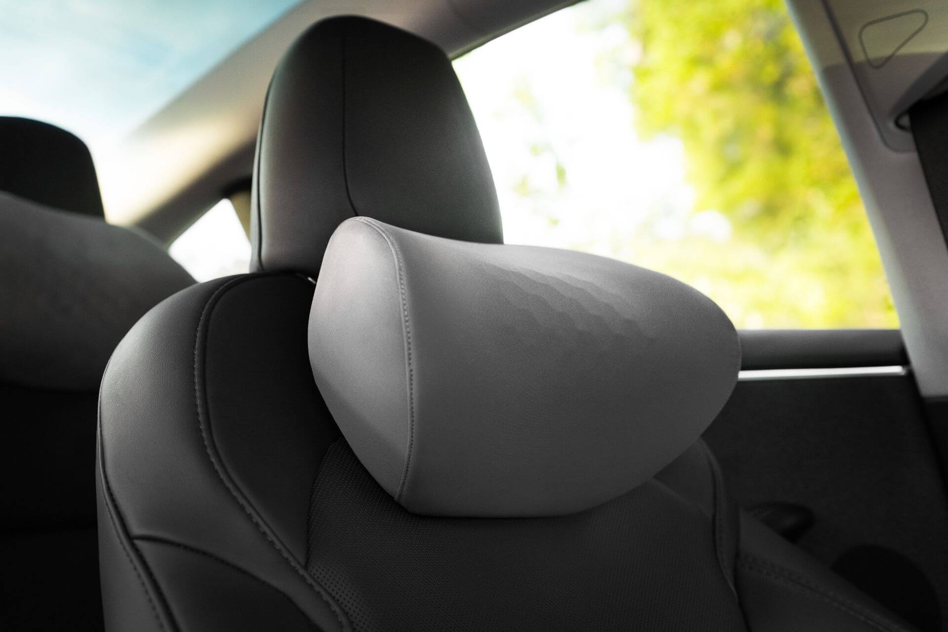 A close-up of the JOWUA Car Neck Pillow displays its ergonomic design and textured surface on a leather headrest. In the background, a blurred view of green foliage through the window hints at an outdoor setting, while the modern interior maintains a sleek appearance.