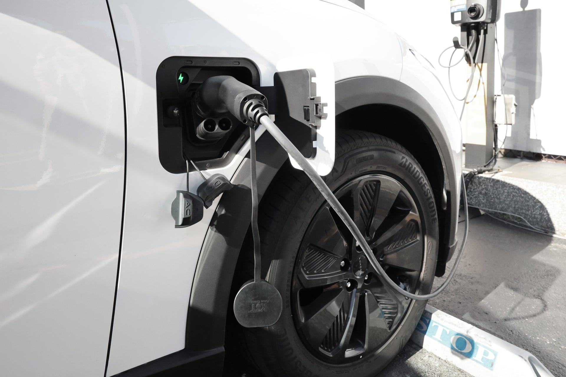 A white electric car featuring black wheels is parked on concrete with a "STOP" marking in the foreground, plugged into a JOWUA CCS2 to CCS1 DC & AC Combined Adapter at a charging port on the side, utilizing DC fast charging.