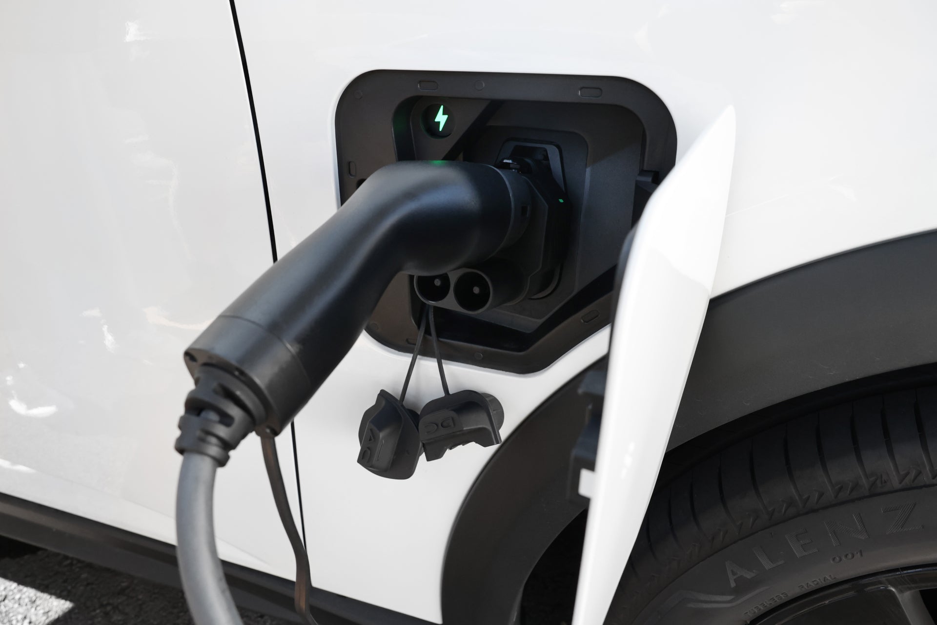 Close-up of an electric car charging with a JOWUA CCS2 to CCS1 DC & AC Combined Adapter. The cable connects the vehicle's side port on the white car, with the cover open and a green light showing DC fast charging.