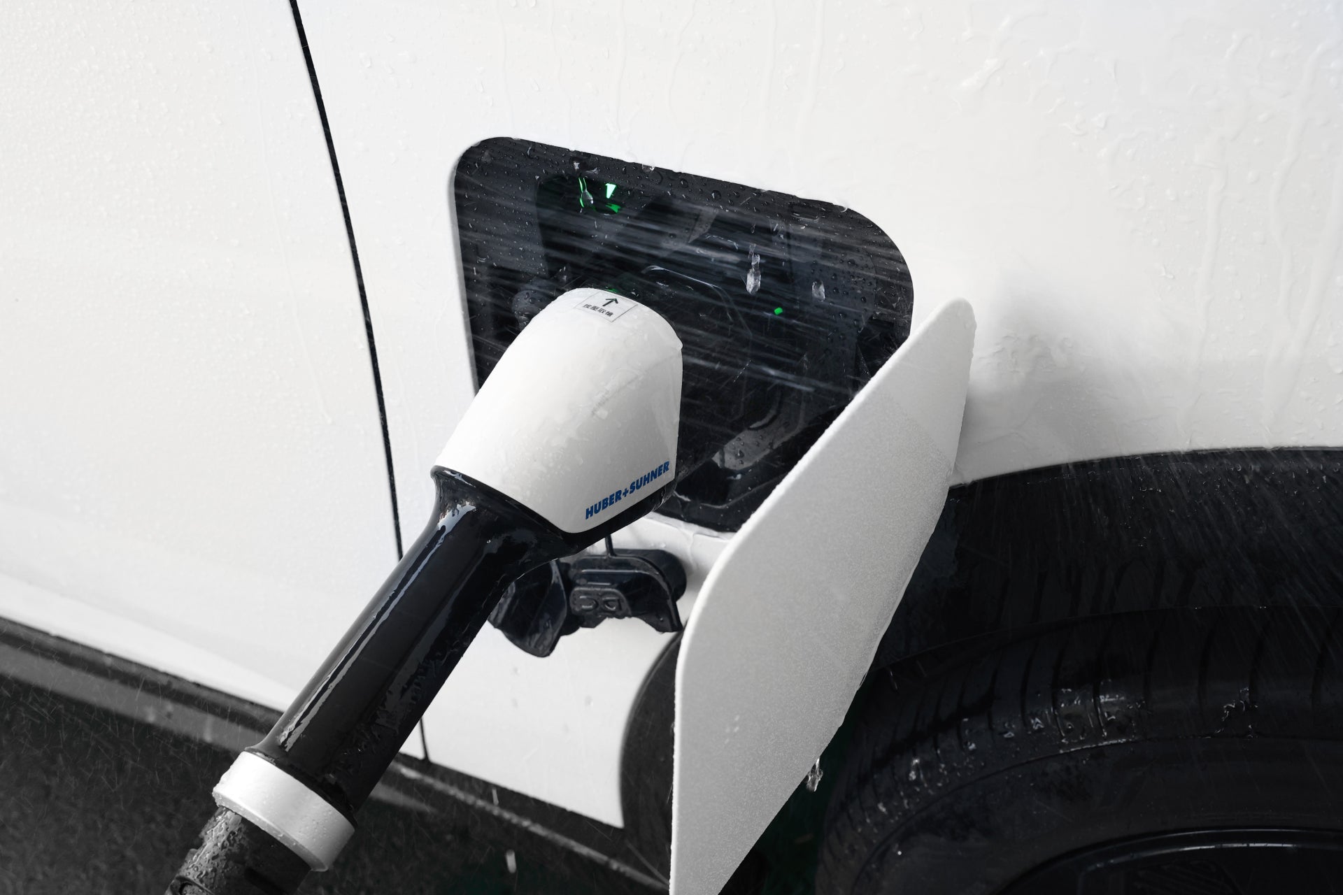 A white electric vehicle charges at a CCS2 station using a JOWUA CCS2 to CCS1 DC & AC Combined Adapter. The cable plugs into the side port for DC fast charging, while rain glistens on the car and surrounding area.