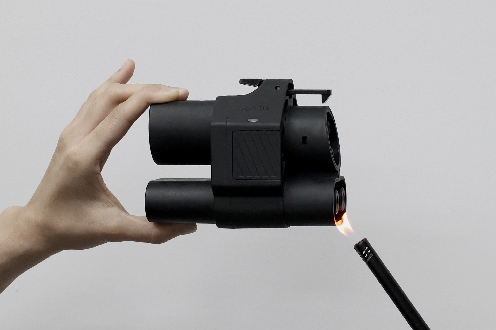 Hands holding a sleek black compact device from JOWUA, the CCS2 to CCS1 DC & AC Combined Adapter, with a lighter igniting a steady flame at the nozzle, reminiscent of the efficiency and power of CCS2 charging stations against a plain white background.