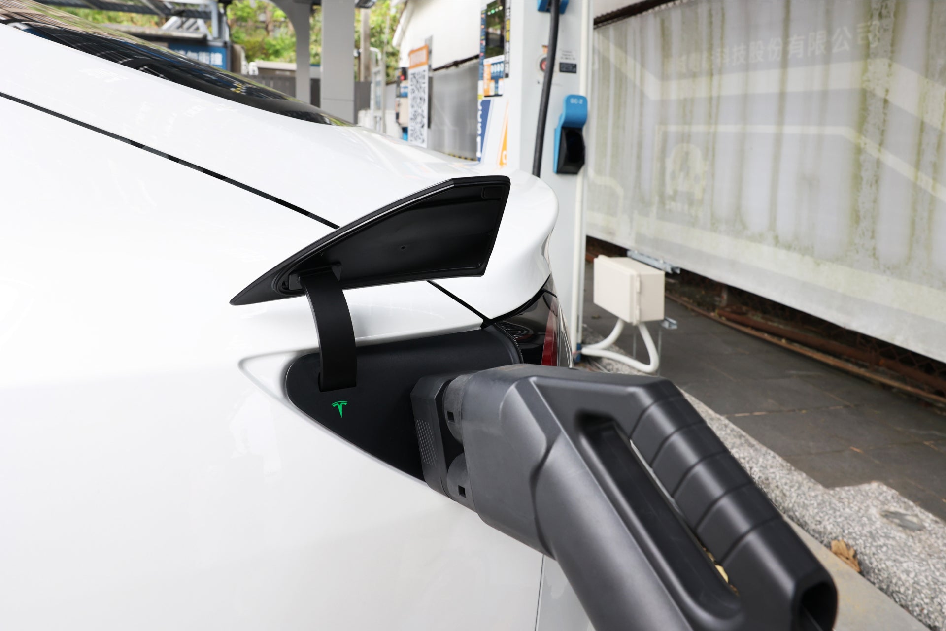 A white electric car is plugged into a charging station using a black JOWUA CCS2 to NACS DC Charging Adapter. The adapter smoothly fits the car's rear port, set against a serene backdrop of greenery and part of a building.