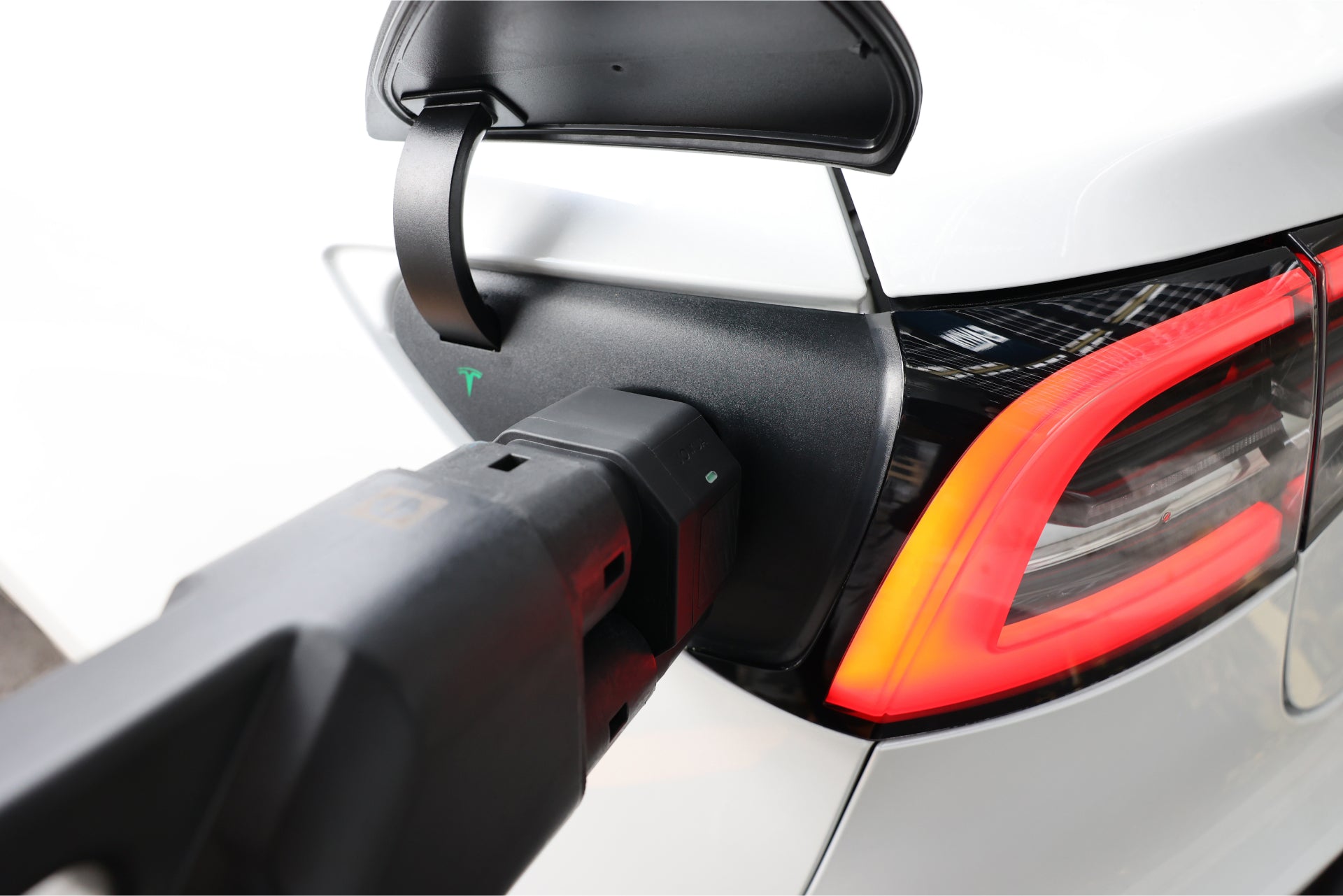 A white electric car is being charged with a JOWUA CCS2 to NACS DC Charging Adapter. The vehicle's charging port is open, and a charger is plugged in while the rear taillight is illuminated.