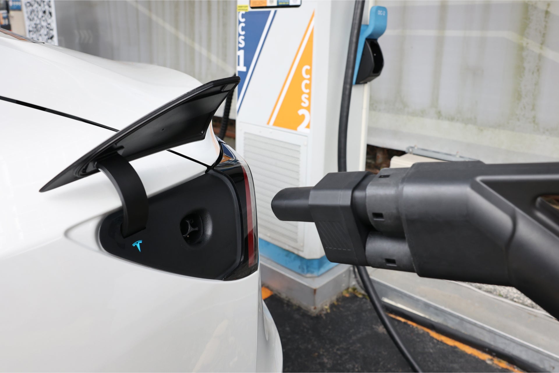 A white electric car is being charged at a station using the JOWUA CCS2 to NACS DC Charging Adapter. The connector is securely plugged into the car's port, with the back of the car and part of the labeled charging station visible, highlighting the DC fast charging process.