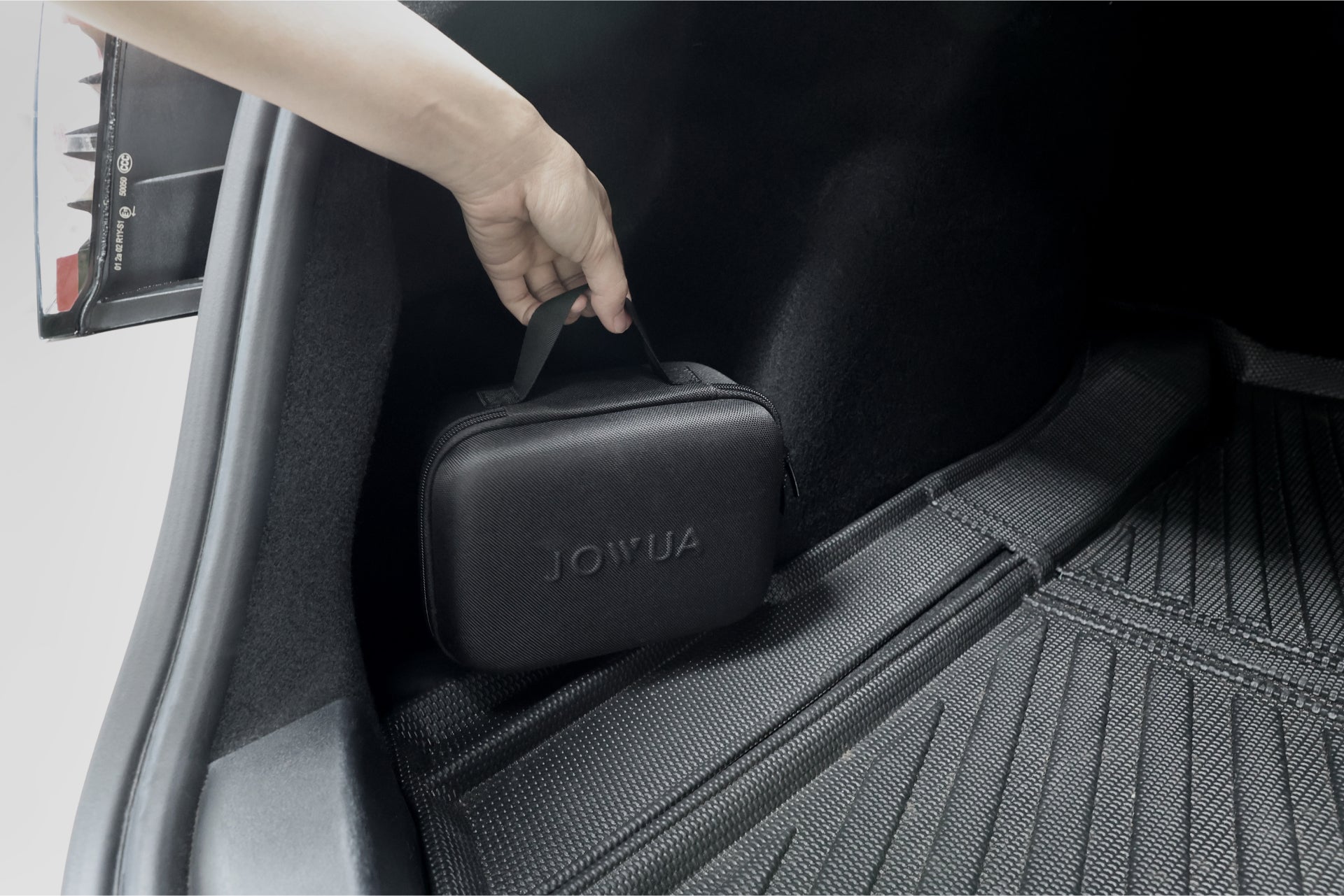 A person places a black JOWUA bag, ideal for carrying items such as the CCS2 to NACS DC Charging Adapter, inside the trunk lined with protective mats. The compact bag features a handle for easy transport.