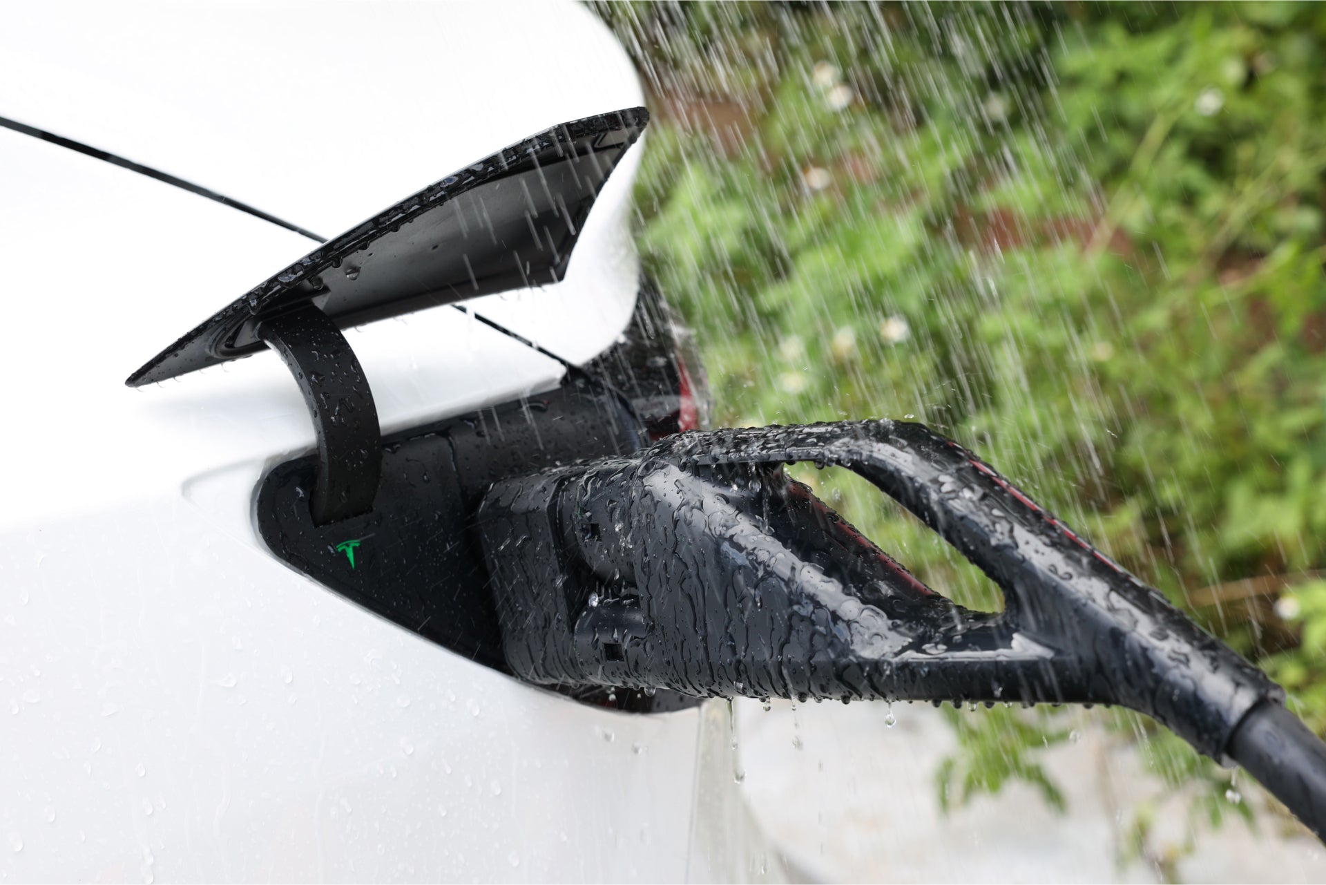 A close-up of an electric car using the JOWUA CCS2 to NACS DC Charging Adapter in the rain shows the charging plug, utilizing the CCS protocol, connected to the vehicle. Water droplets glisten on its surface as green foliage softly blurs in the background.