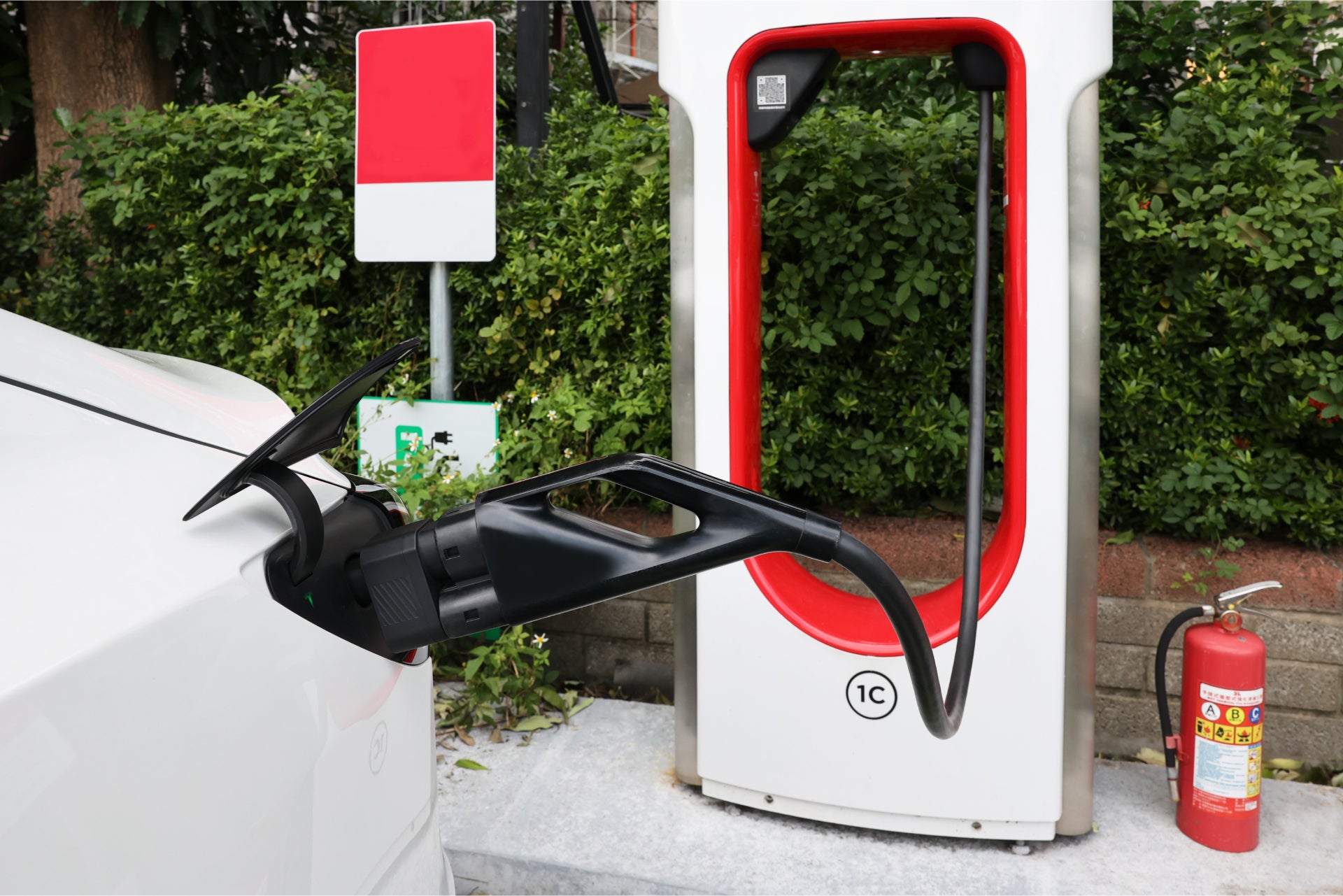 A white car uses the JOWUA CCS2 to NACS DC Charging Adapter at a station outlined in red, near a red and white fire extinguisher, against green bushes, highlighting the seamless integration of sustainable technology.