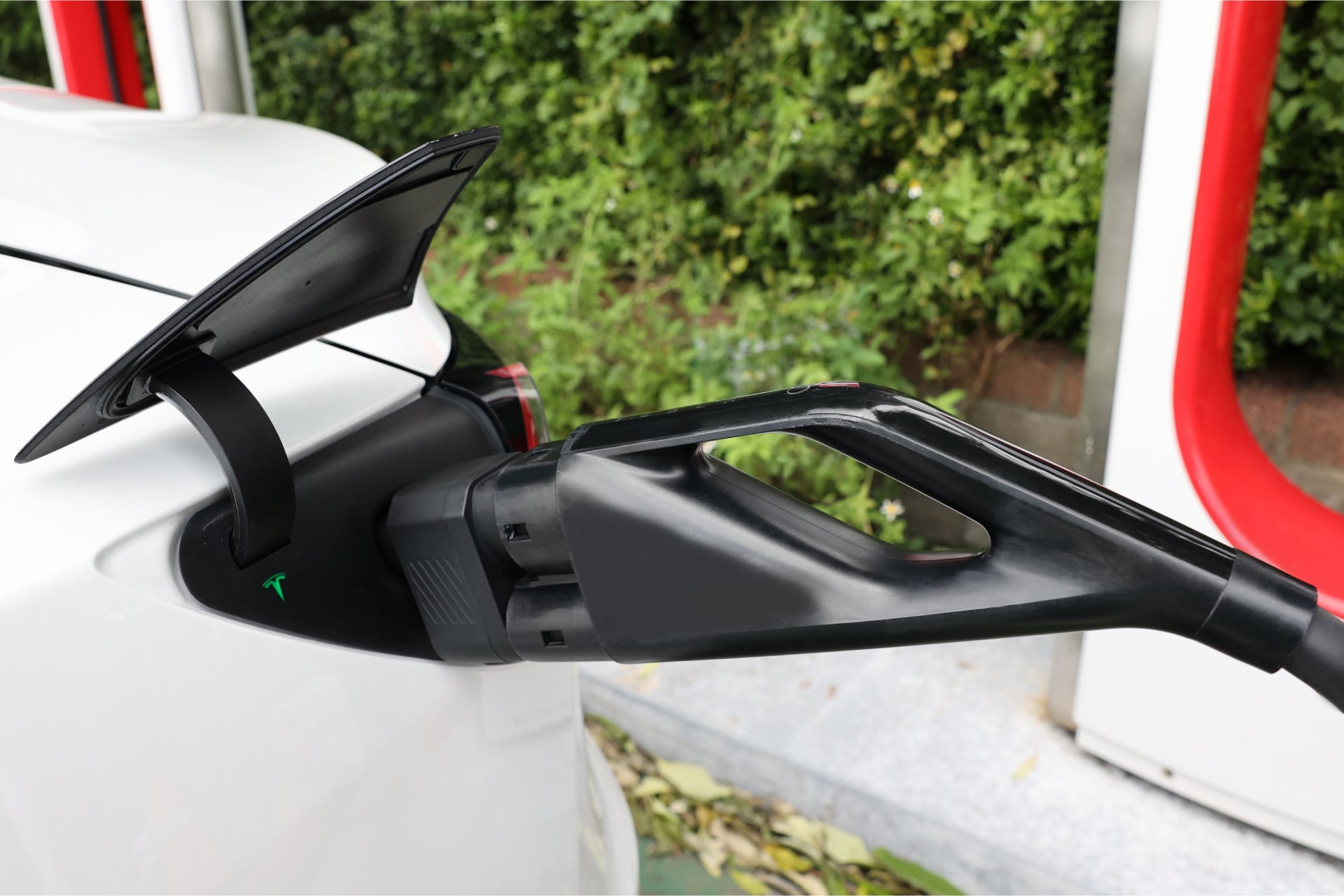 A close-up shows a white electric car being charged with a JOWUA CCS2 to NACS DC Charging Adapter, the cable plugged in and port cover open. Greenery in the background offers a serene contrast to the advanced technology.