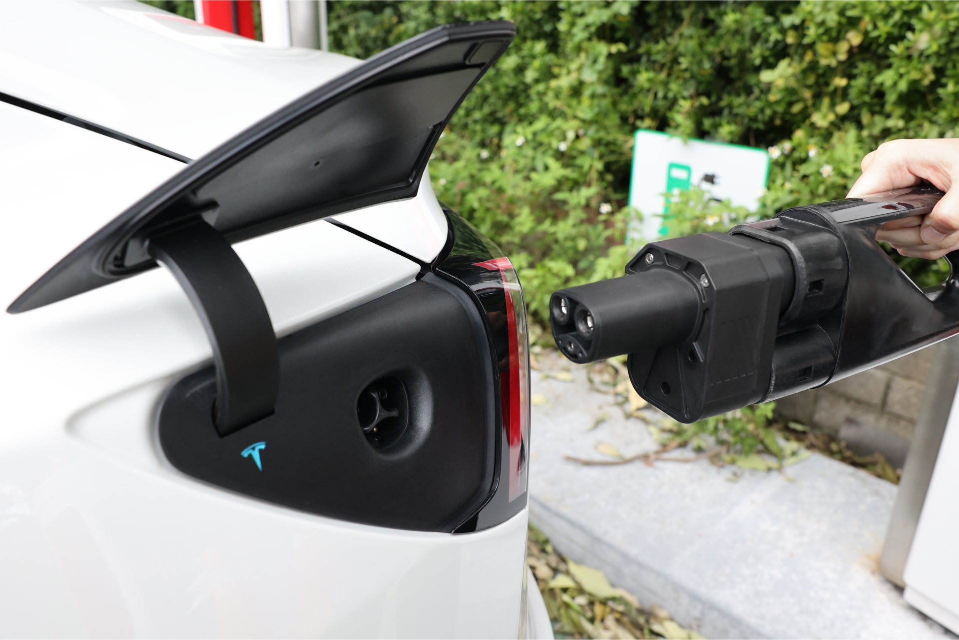 A person holds a JOWUA CCS2 to NACS DC Charging Adapter near the open charging port of a white electric car parked by greenery.