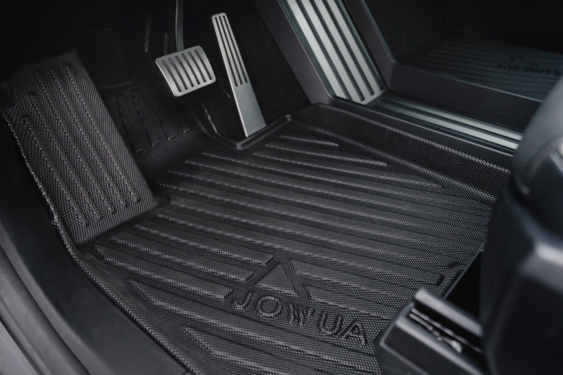 Close-up of a car interior highlights a textured black JOWUA All Weather Floor Liner in the driver's footwell, with embossed branding. The metal pedals and door sill evoke the durability of a Tesla Cybertruck, ideal for all-weather protection.