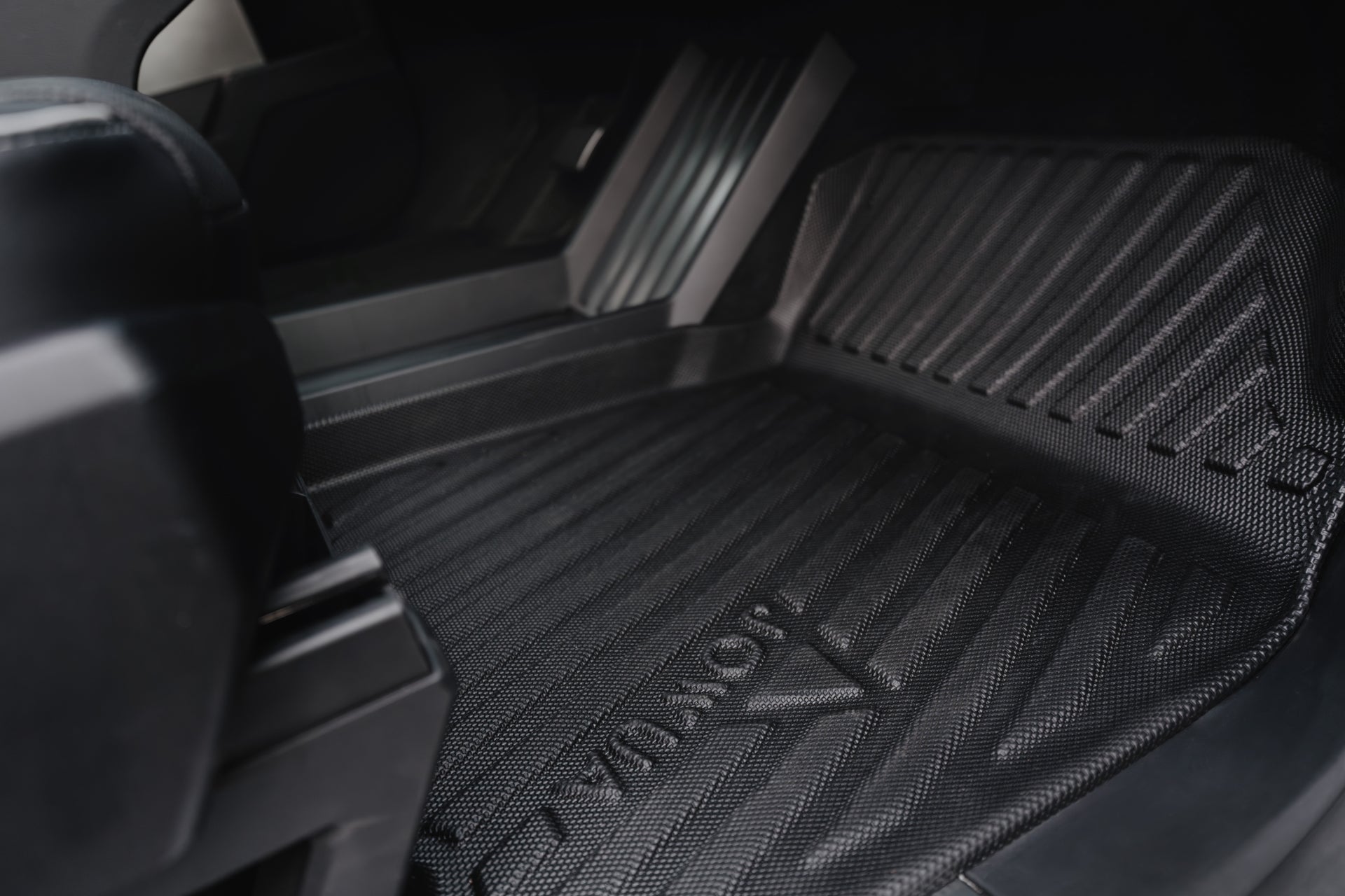 Close-up of a Tesla Cybertruck's interior footwell features a dark, rigid all-weather floor liner from JOWUA with grooves and "JOWUA" embossed on it, offering superb protection against dirt and moisture.