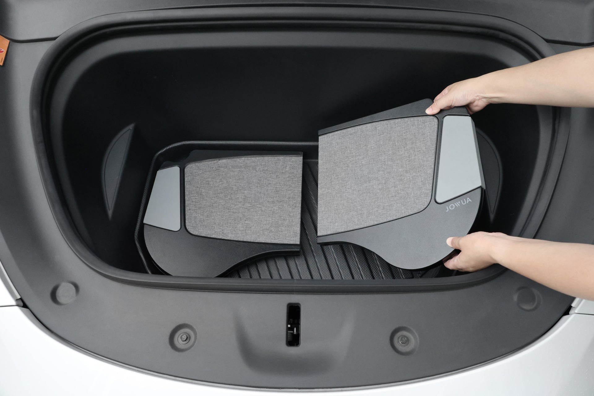 Hands placing a set of gray and black JOYJUA protective floor mats into the trunk of a Tesla Model S. Crafted from premium materials, these mats feature a sleek design that fits neatly into the space.