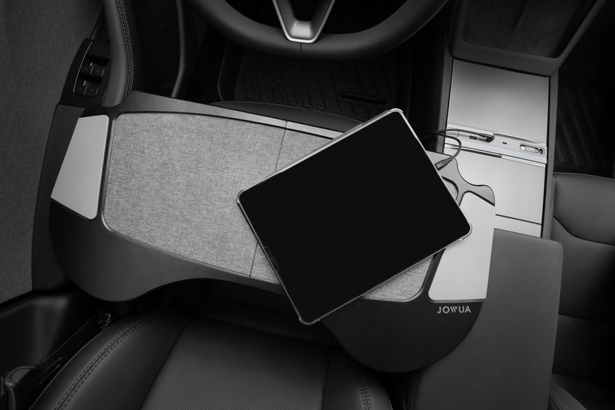 An overhead view highlights a stylish steering wheel and center console in the interior, accompanied by a JOWUA Foldable Car Tray Bundle (Reengineered Model 3 Highland, 2024+) fitted over the driver's seat. A tablet rests on this black and gray lap desk crafted from PU leather, which complements its sleek, modern appearance.