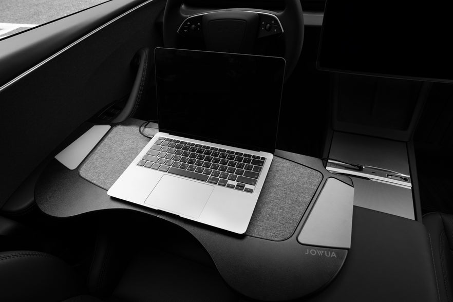 A laptop sits on the JOWUA Foldable Car Tray Bundle, tailored for the Model 3 Highland (2024+) Tesla interior. The steering wheel and part of the dashboard are visible, while the tray, crafted with PU leather, offers a cushioned surface and a flat workspace inside the vehicle.