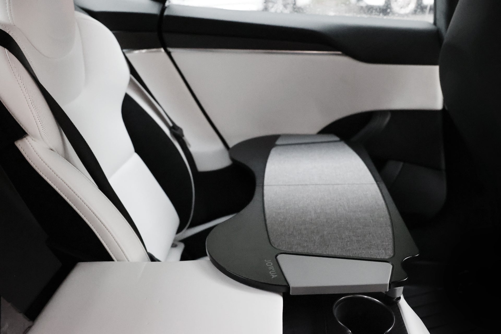 The interior of this Tesla car boasts a sleek, modern design, featuring white seats with black accents made from premium PU leather. Attached to the passenger seat is the JOWUA Foldable Car Tray, which includes a convenient cup holder. This accessory is ideal for enhancing comfort while enjoying views through the ample windows of your Model 3.