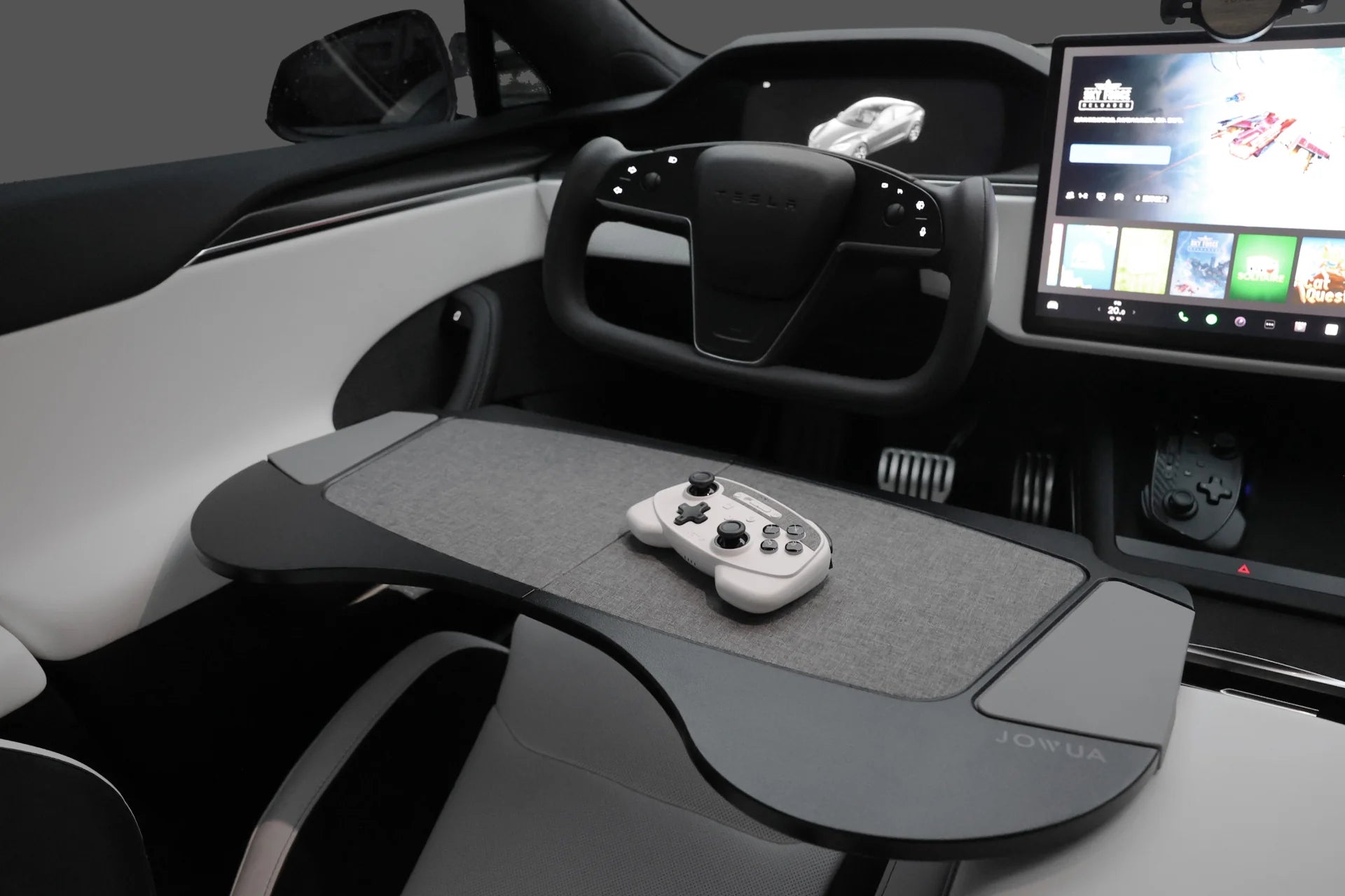 The interior of this car boasts a modern steering wheel and dashboard, highlighted by a digital display for games. A gaming controller is conveniently situated on a foldable car tray attached to the driver’s seat, creating an exceptional gaming setup with Tesla Christmas: The Ultimate Bundle from JOWUA enhancing your drive.