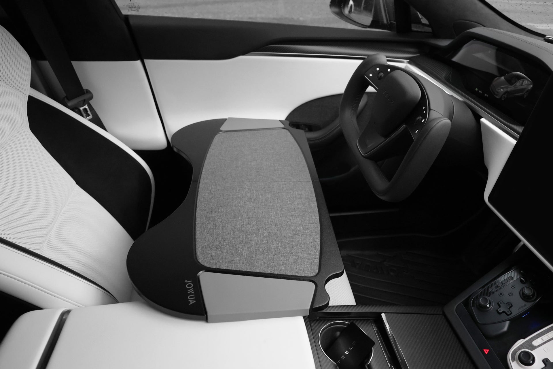 The interior of this modern vehicle features a distinctive steering wheel design, a large central touchscreen, and a sleek dashboard. For an added touch of convenience, the driver's seat is equipped with JOWUA's Foldable Car Tray made from premium PU leather, making it the perfect accessory for fans of the Tesla Model S/3/X/Y.