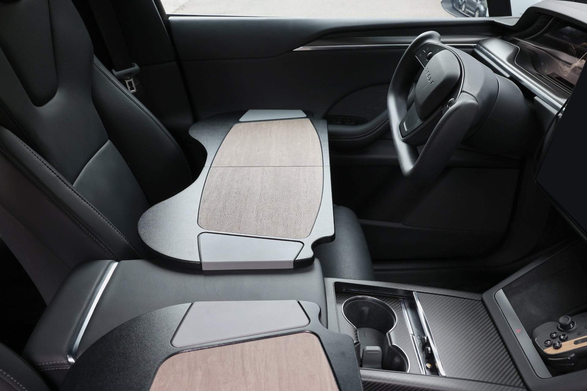 The interior of a contemporary vehicle, enhanced by JOWUA's Foldable Car Tray for Tesla, exudes stylish black and wood accents. It includes spaciously designed tables above the passenger seats crafted from high-quality materials and a dashboard equipped with a digital display.