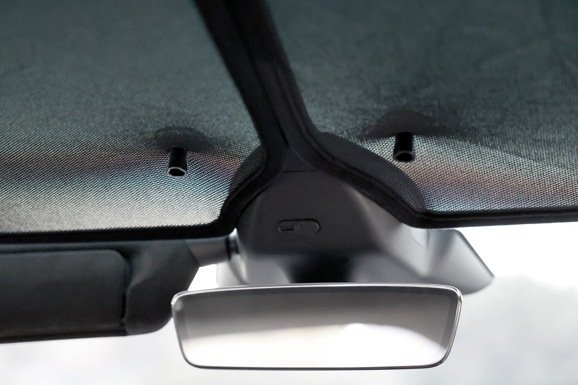 Close-up of a car's rearview mirror and JOWUA Glass Roof Sunshade-Mesh (Model X) sun visors inside a vehicle. The visors, providing UV protection, are folded up, with a button near the mirror visible from the driver's seat perspective.