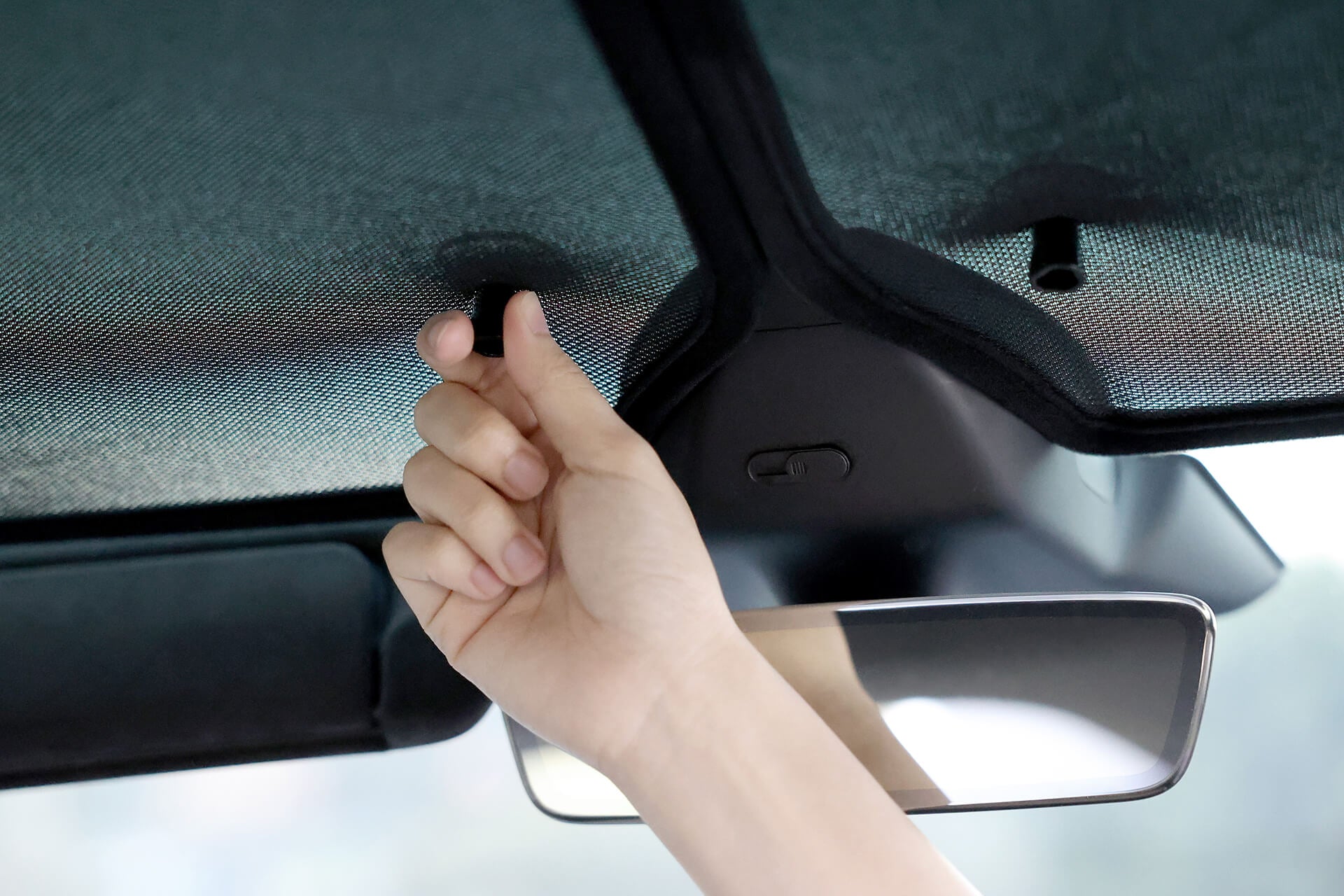 A hand pulls down the JOWUA Mesh Glass Roof Sunshade made of UV protection fabric from the car roof interior, with the rearview mirror visible in the foreground, enhancing comfort and style under the Model X sunroof.