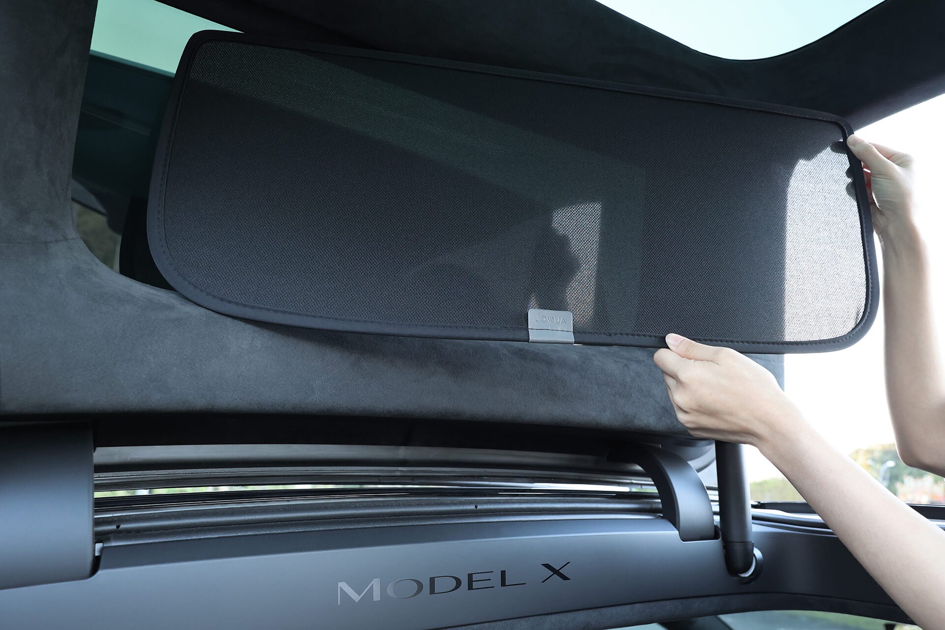 A person meticulously installs the JOWUA Model X Separate Sunshade Bundle on their Tesla Model X's panoramic windshield, adjusting the mesh screen for optimal heat insulation, while the iconic Model X logo shines on the door frame.