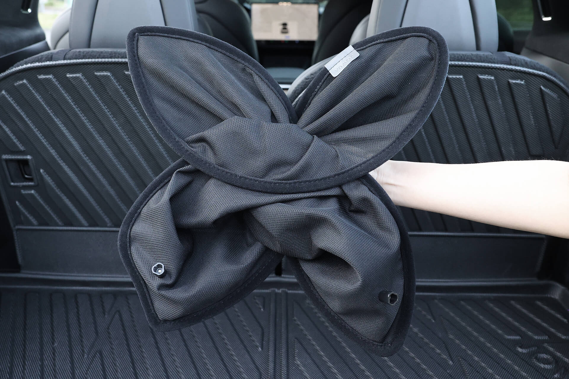 A hand holds a folded JOWUA Glass Roof Sunshade - Separate Design (Model X) in the trunk, showcasing its flower shape and UV-blocking design. The black interior with folded seats highlights its practical and stylish heat insulation.