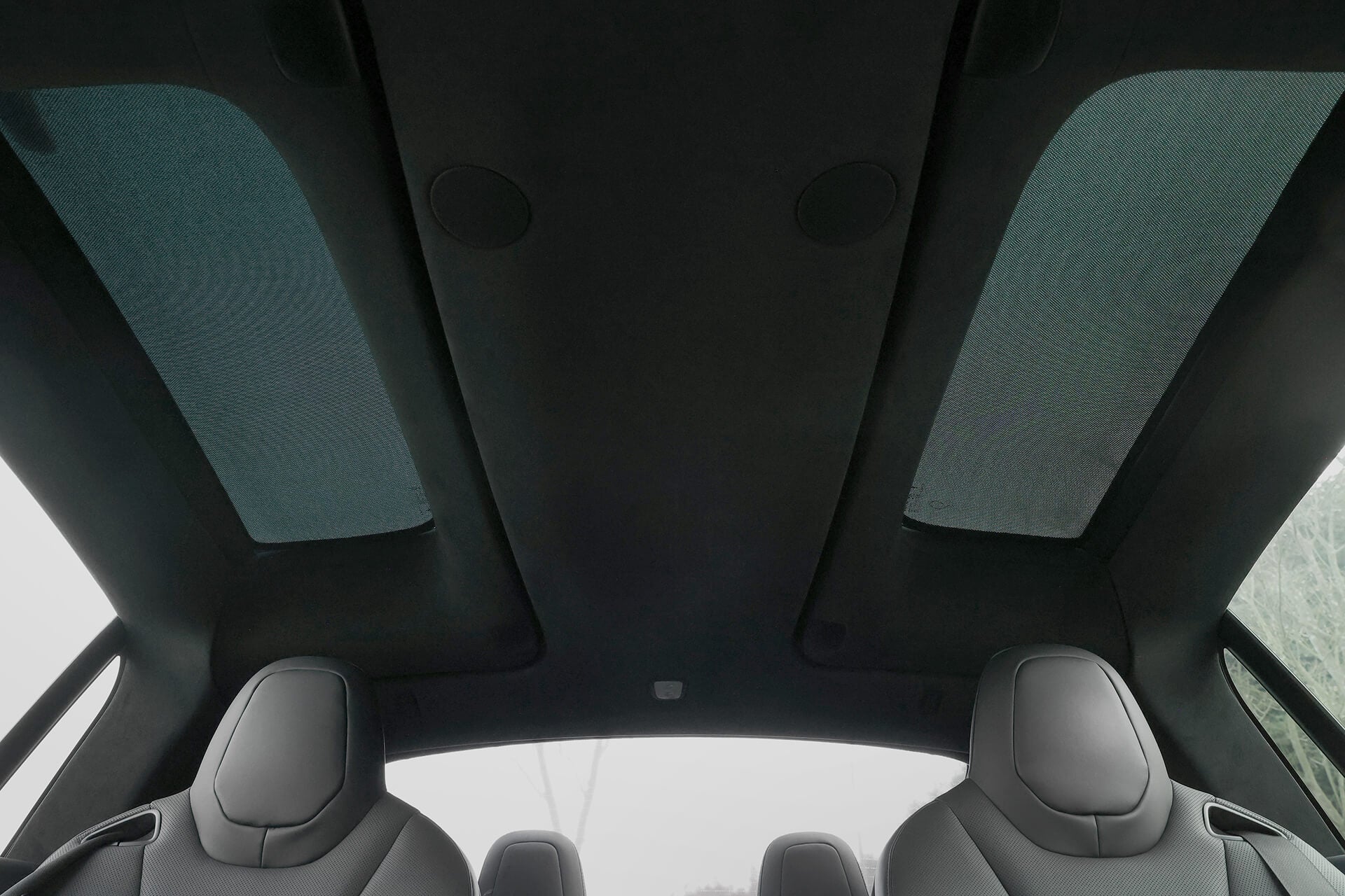 Interior view of a car highlighting its JOWUA Glass Roof Sunshade-Mesh (Model X) with sleek design. Visible are two front seats with headrests and the top of a back seat, all under elongated glass panels providing UV protection and natural light.