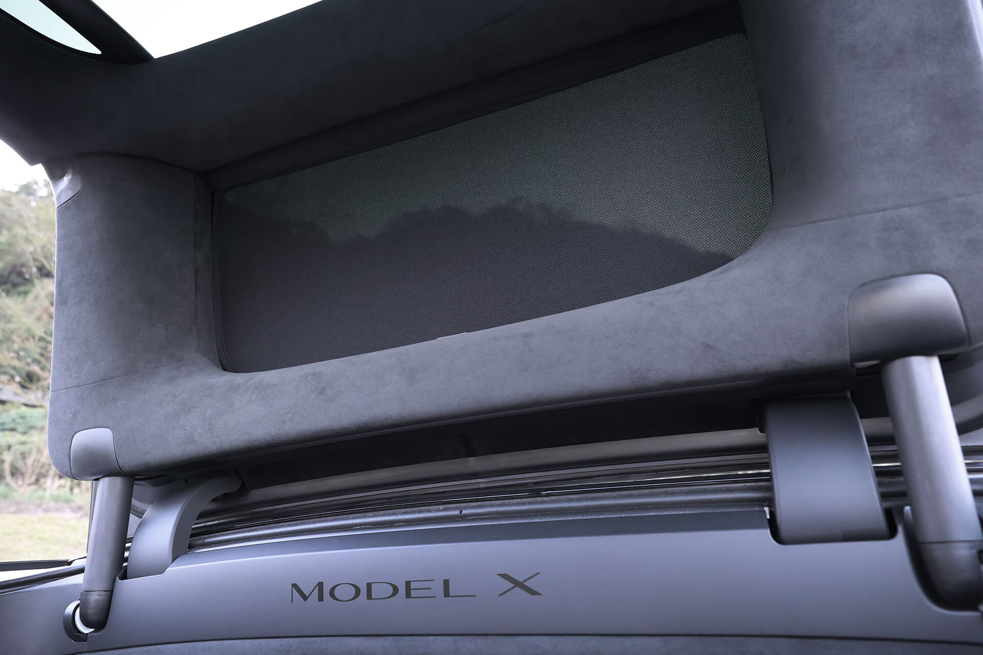 A car interior view shows a storage compartment above the rear seats, with "MODEL X" elegantly displayed on the trim beneath. The area has dark upholstery and features the JOWUA Glass Roof Sunshade-Mesh (Model X) for added UV protection.