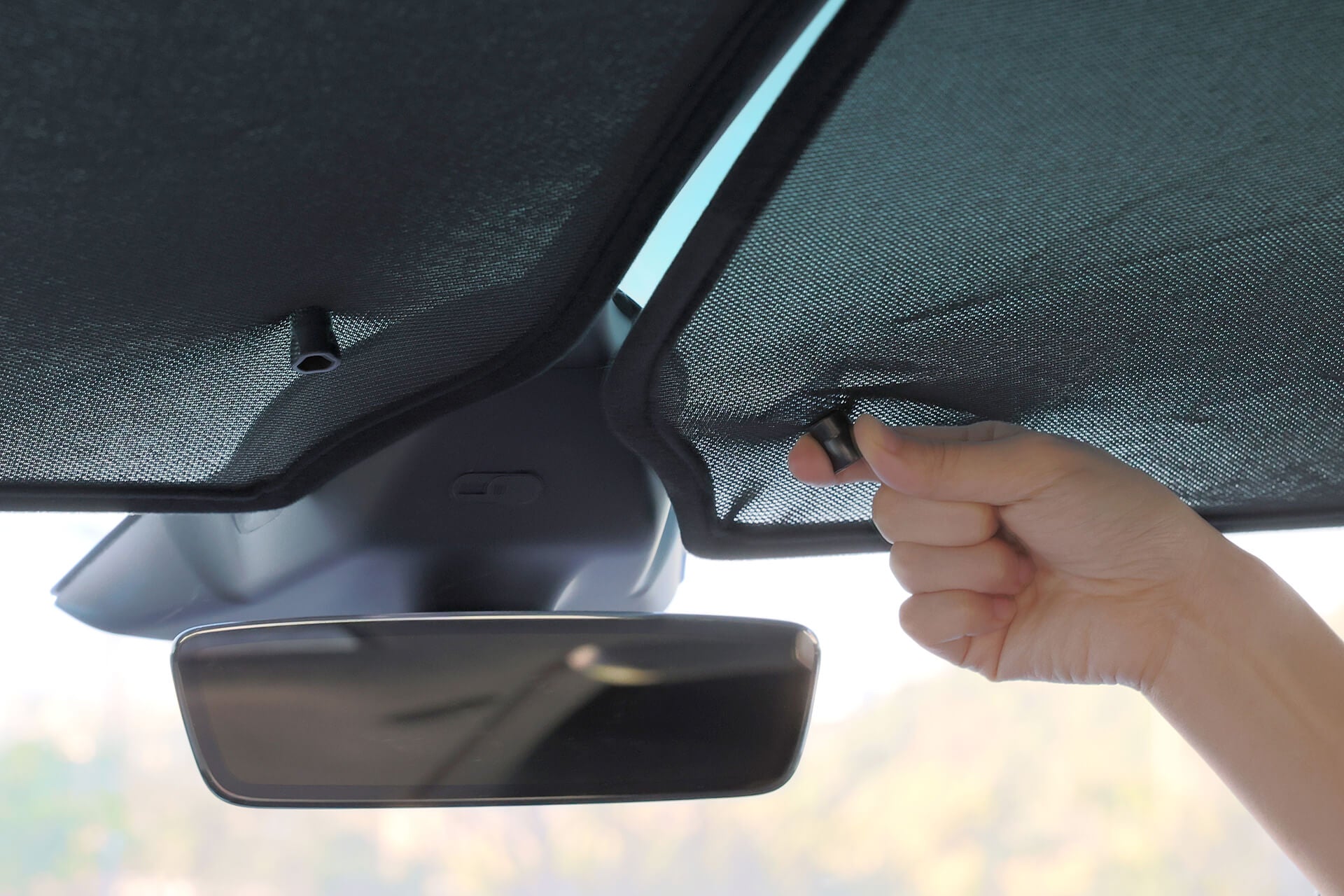 A person adjusts the JOWUA Glass Roof Sunshade- Separate Design (Model X) inside the car, pulling down a fabric screen for UV protection. Below the sunshade, the rearview mirror is visible against a sunny, blurred background.