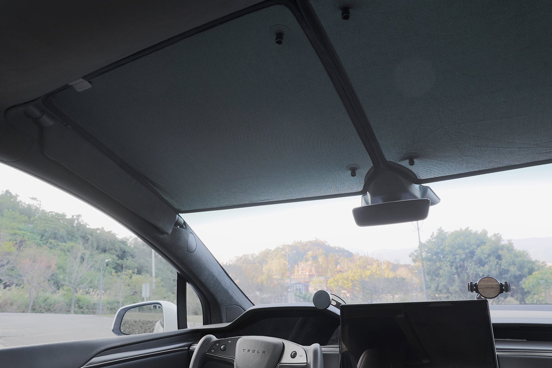 The car's interior showcases a JOWUA Glass Roof Sunshade-Mesh (Model X) for UV protection. The dashboard includes a steering wheel and digital display, while scenic trees and mountains are visible through the windshield. Heat insulation maintains cabin coolness.